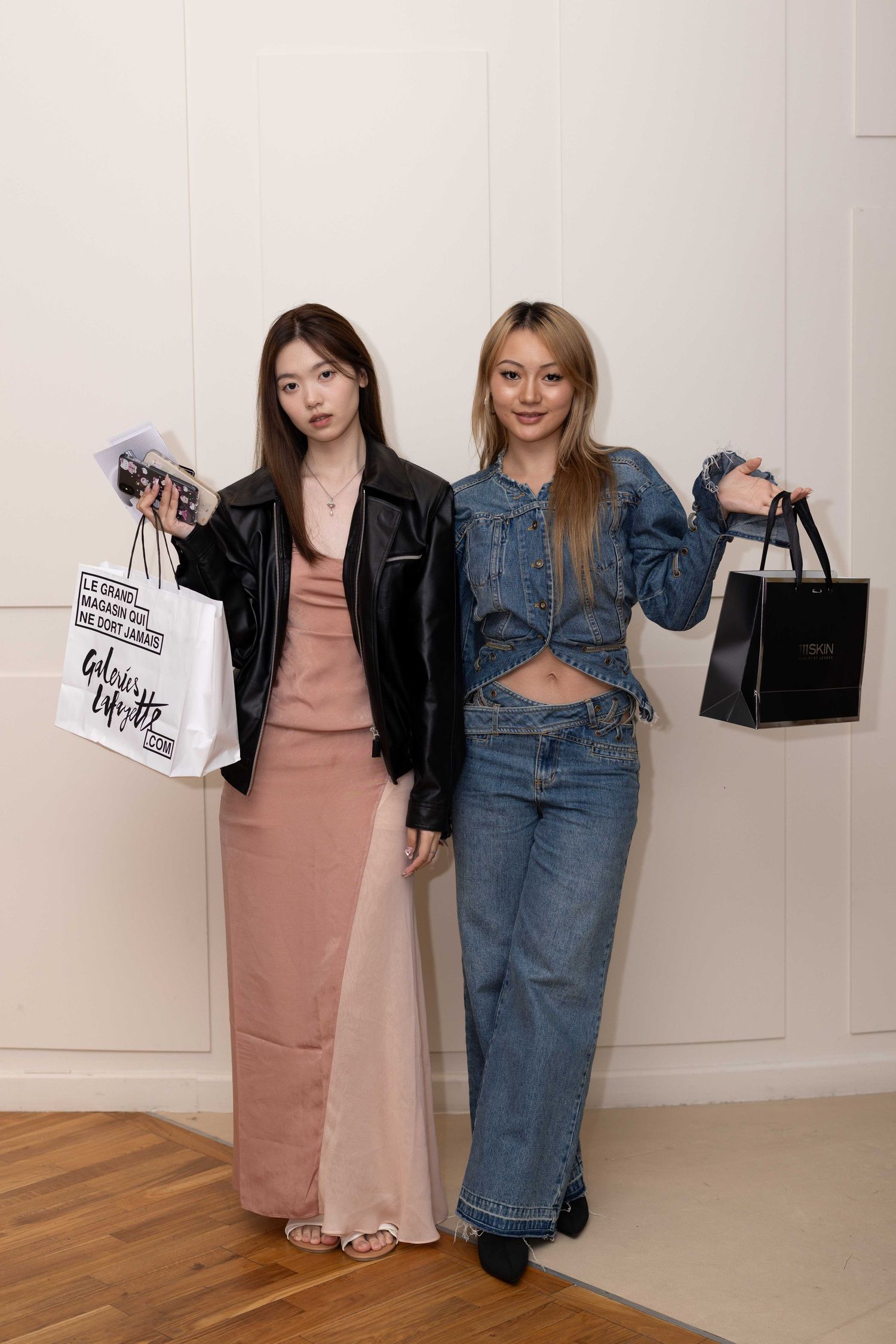 Two friends in casual outfits showing off their shopping bags against white background.
