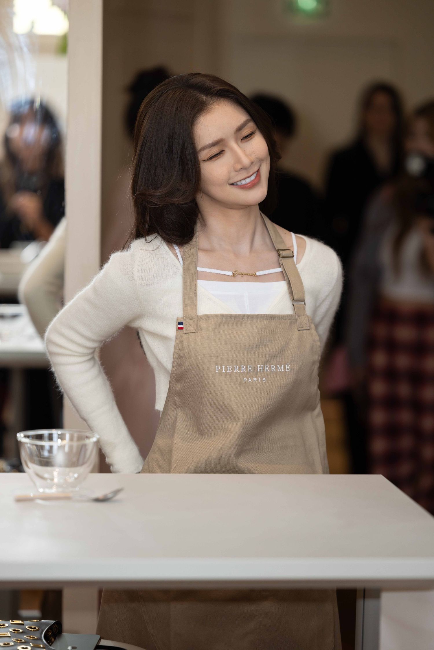 Person in beige apron and white sweater seated at white counter during event.