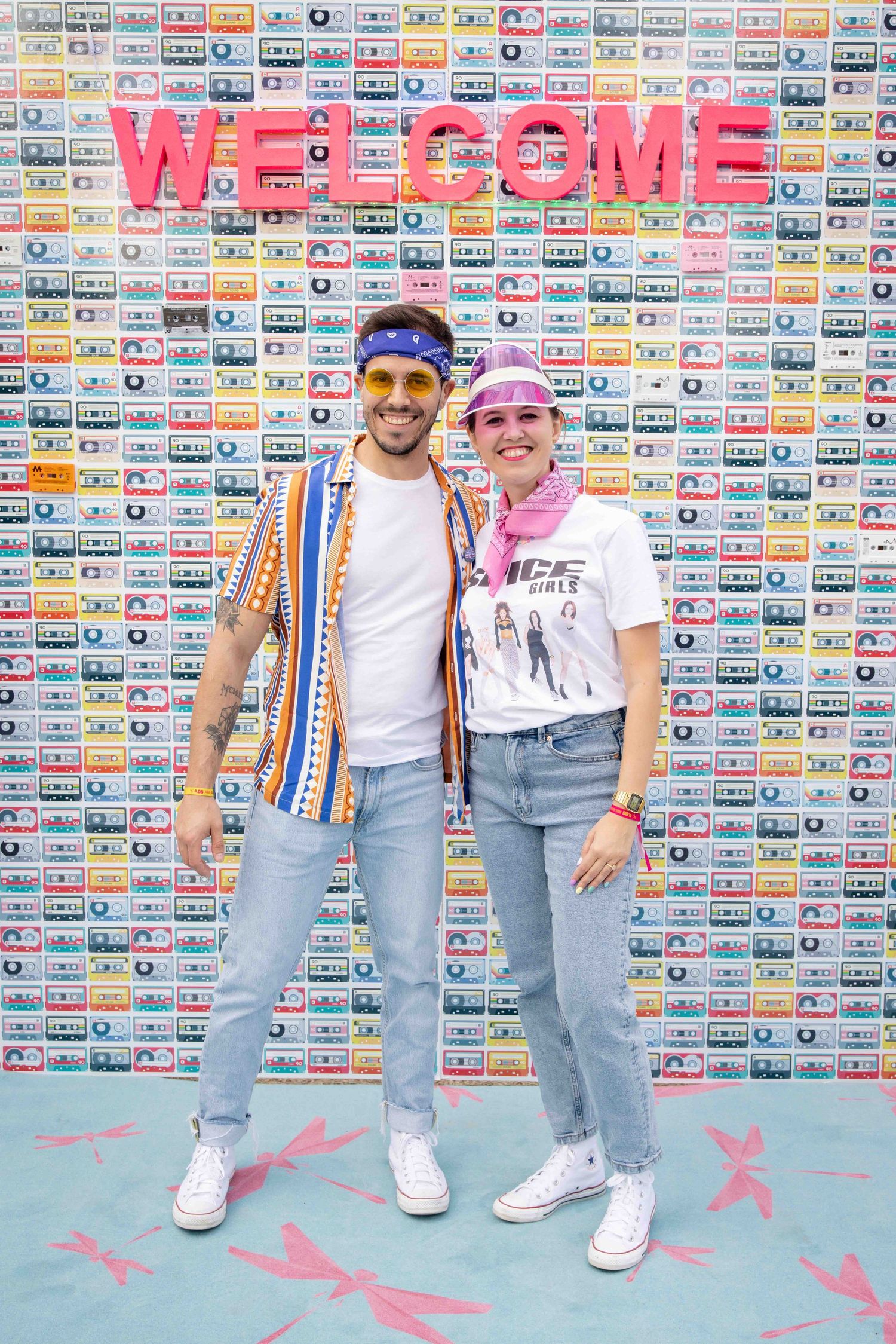 Group wearing denim and colorful accessories poses in front of wall covered in retro cassette tapes.