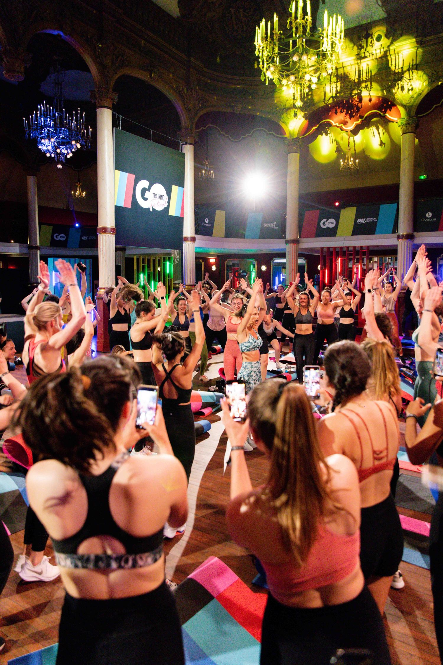 Large group of fitness enthusiasts with raised arms during an energetic workout class in a dimly lit venue.