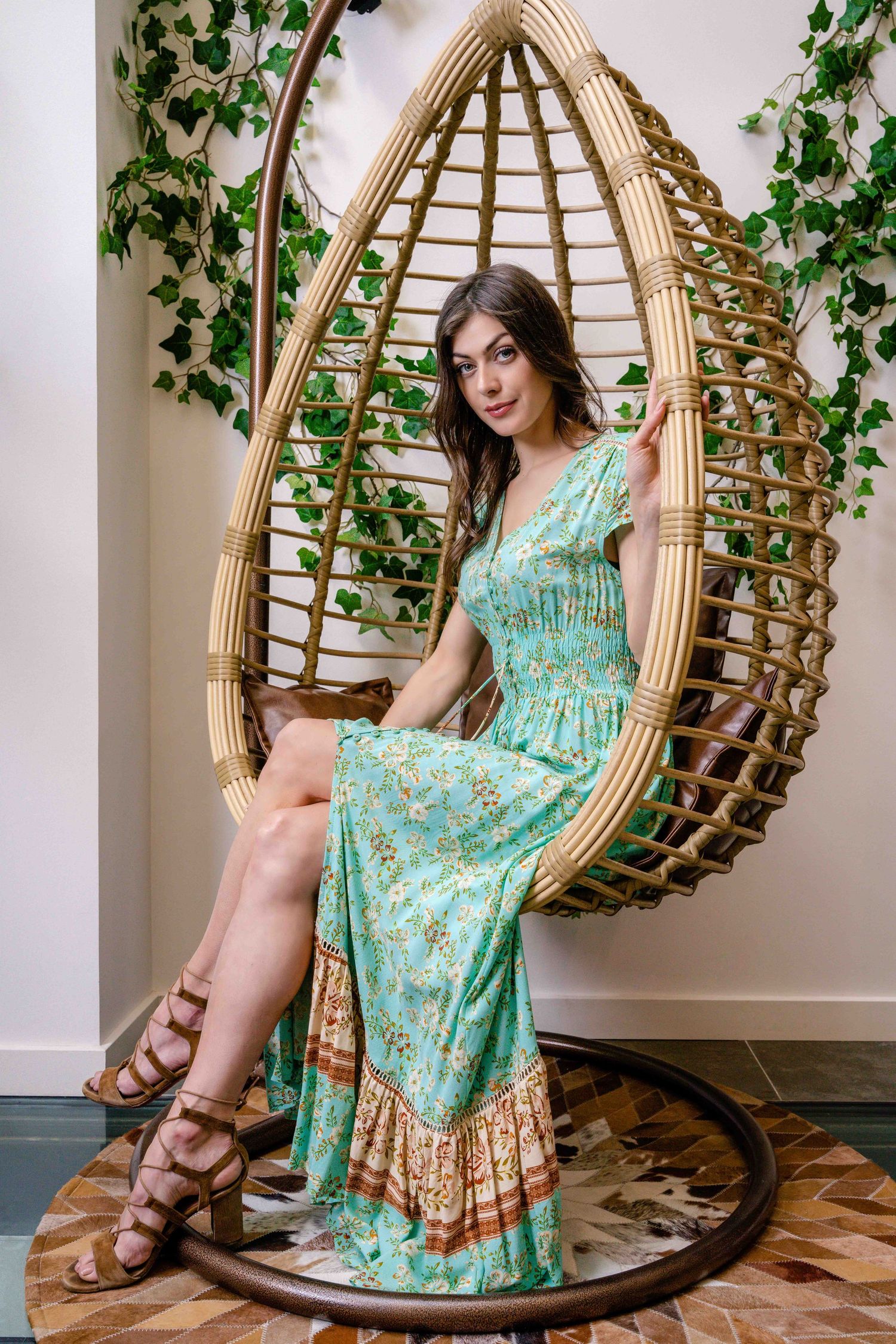 Le mannequin se détend dans un fauteuil suspendu en rotin et porte une robe longue fleurie turquoise.