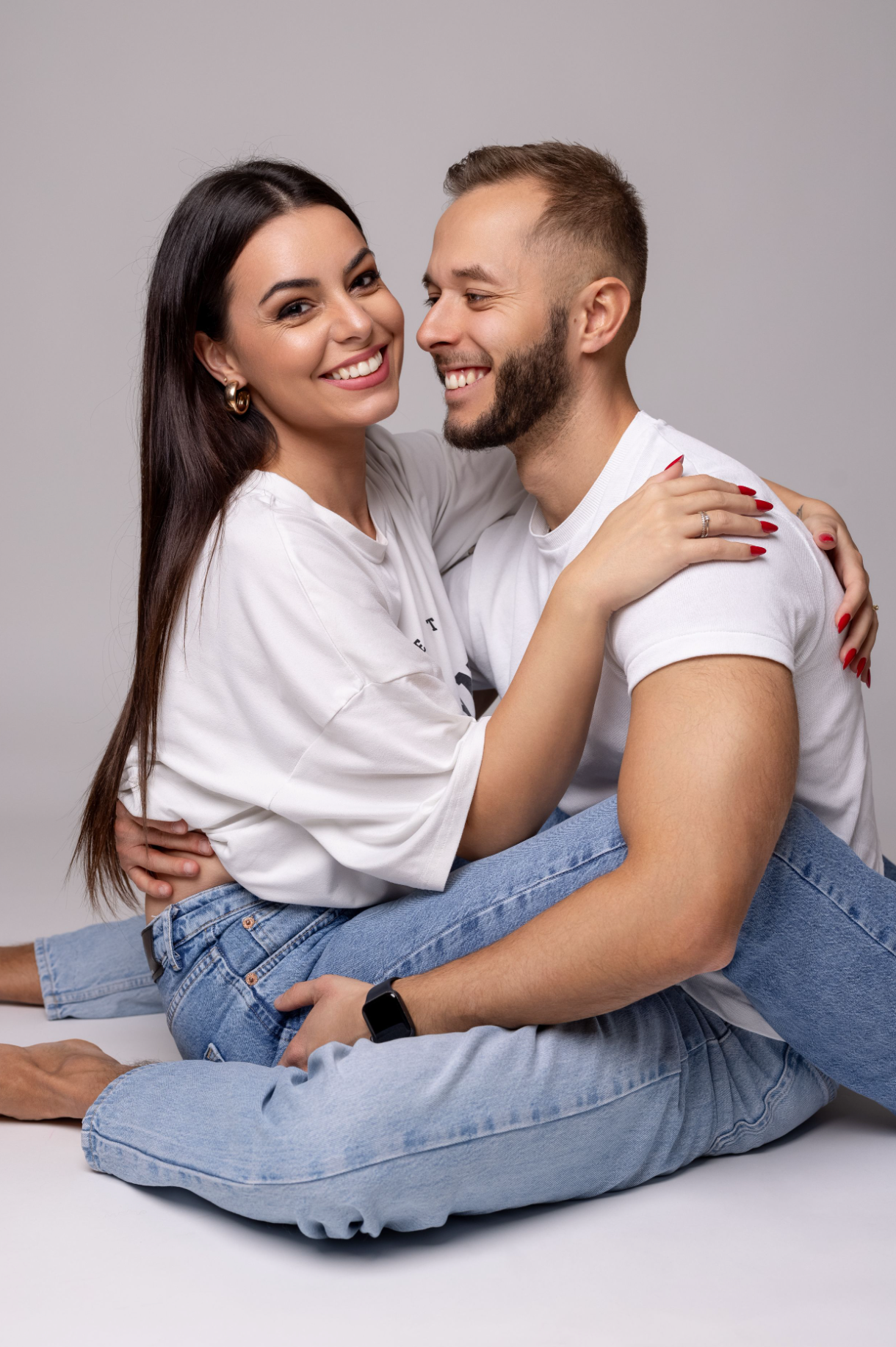 A couple in matching white shirts and jeans embrace and laugh together while sitting on a light gray background.