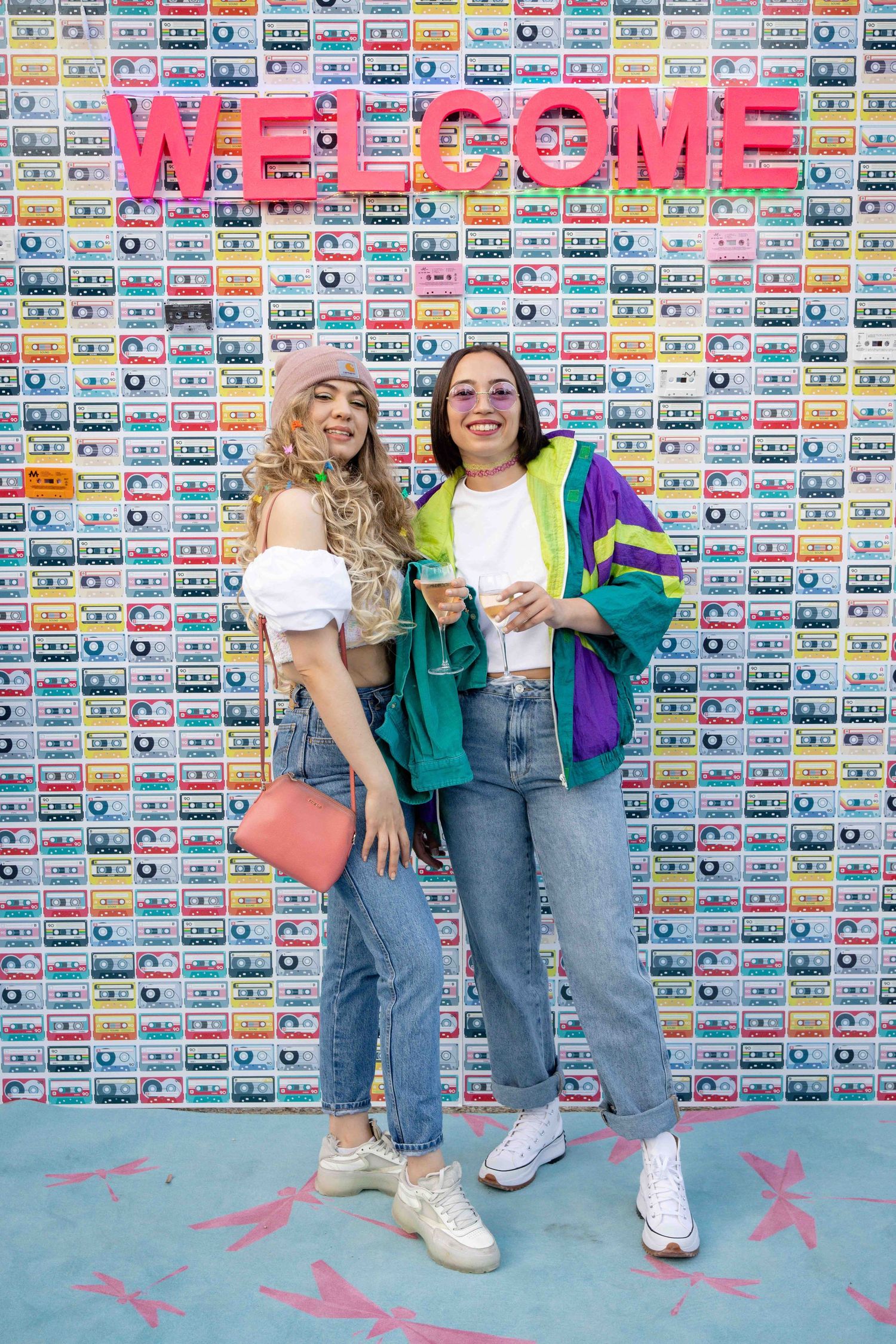 Two friends in trendy outfits pose together against a wall decorated with retro cassette tapes.