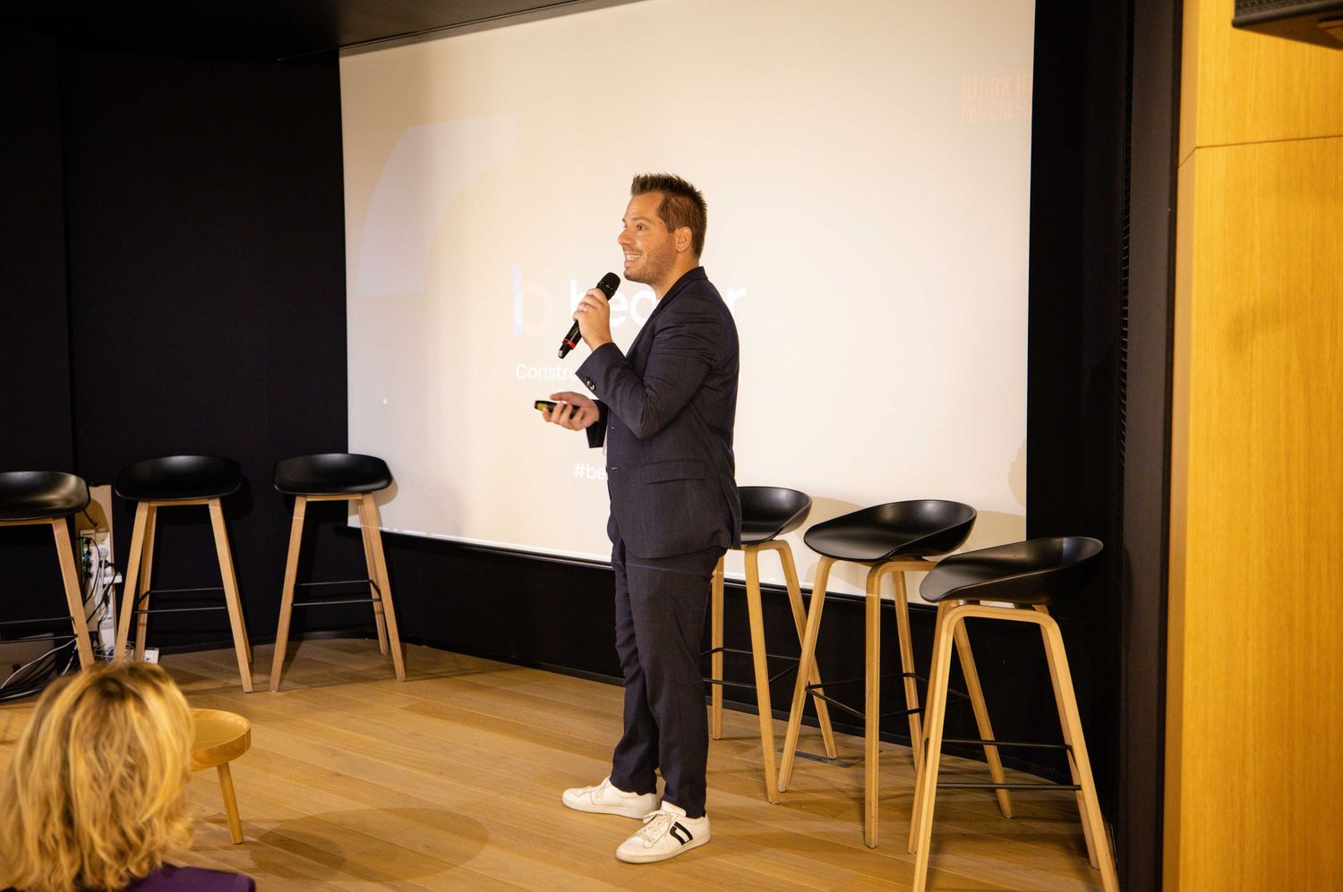 Presenter stands on stage with projection screen displaying presentation content.