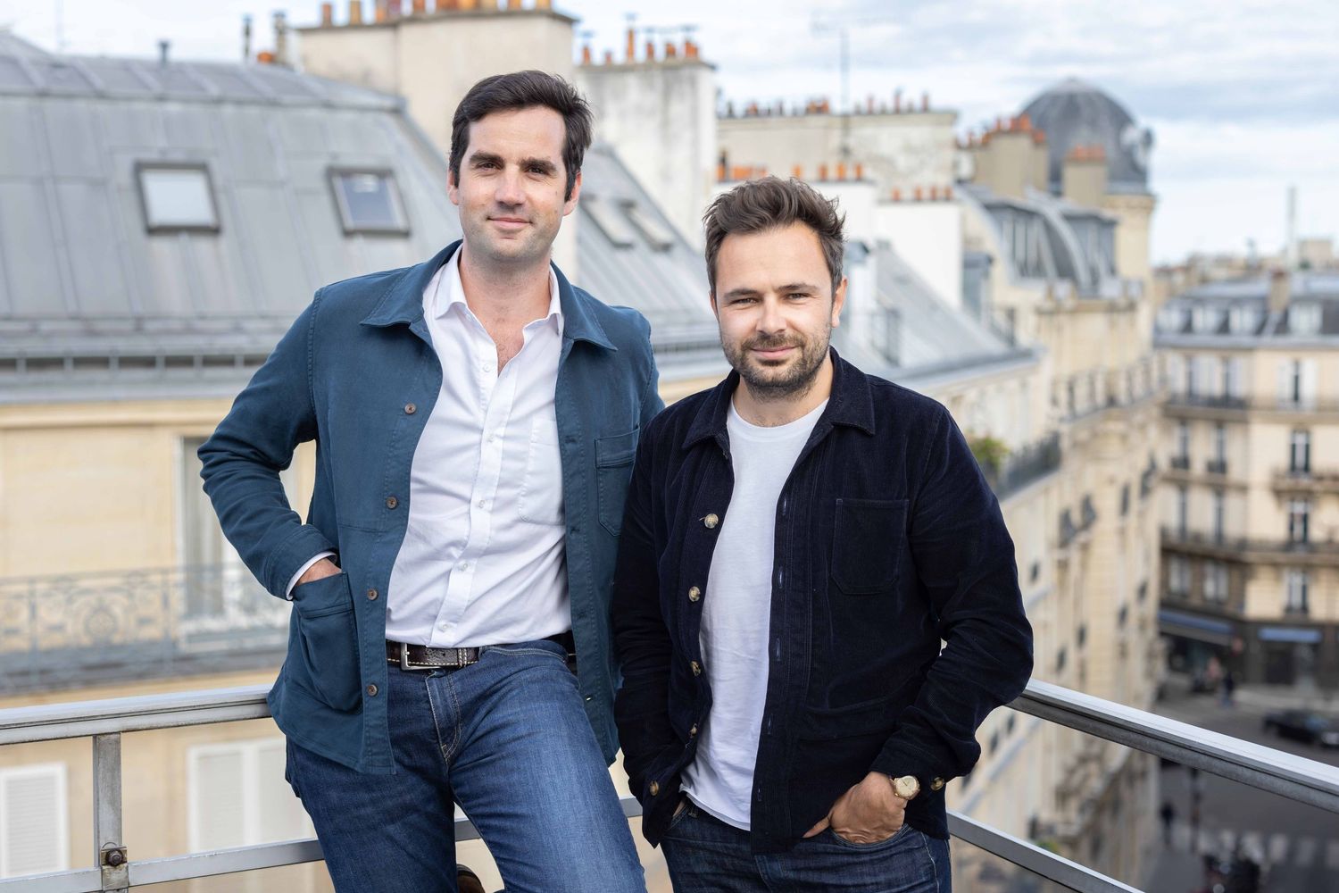 Two people posing casually on a balcony with Parisian architecture behind them.