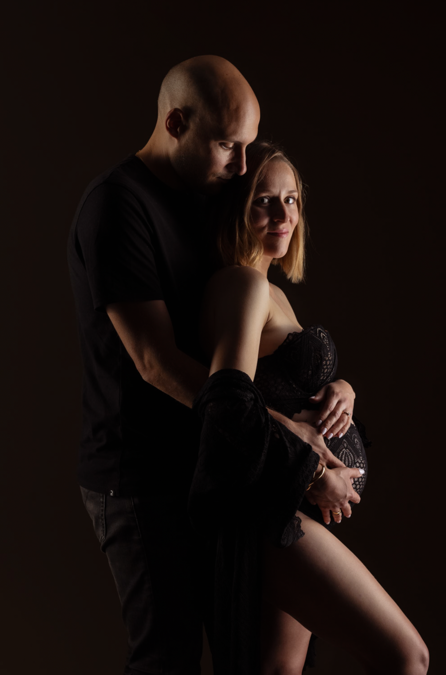 Intimate low-key portrait of a couple embracing in dramatic lighting against a dark background.