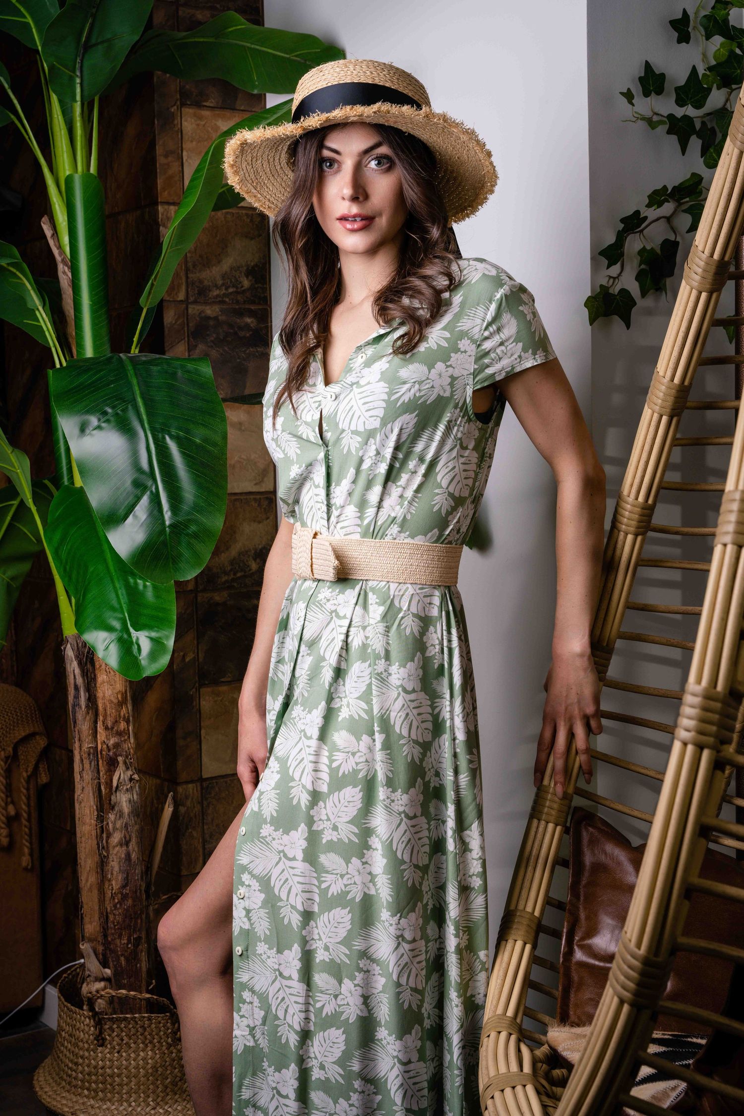 Un mannequin en robe fleurie verte de style vintage pose avec des meubles en bambou et des plantes tropicales.