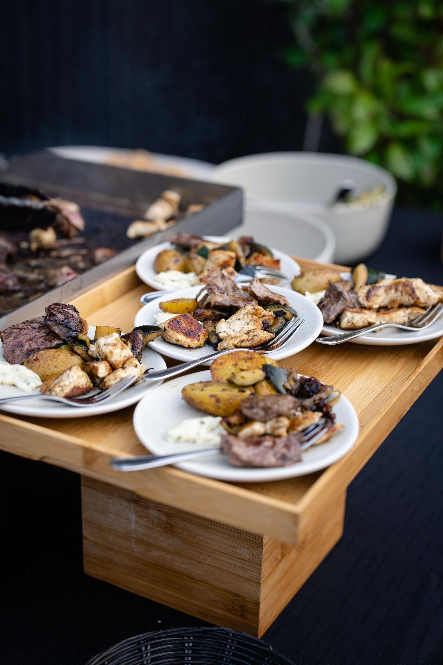 Grilled meats and sides plated on white dishes displayed on a wooden serving board.