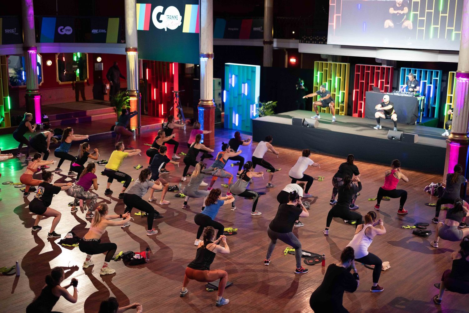 Large group fitness class with colorful lighting doing synchronized exercises in a modern studio space.