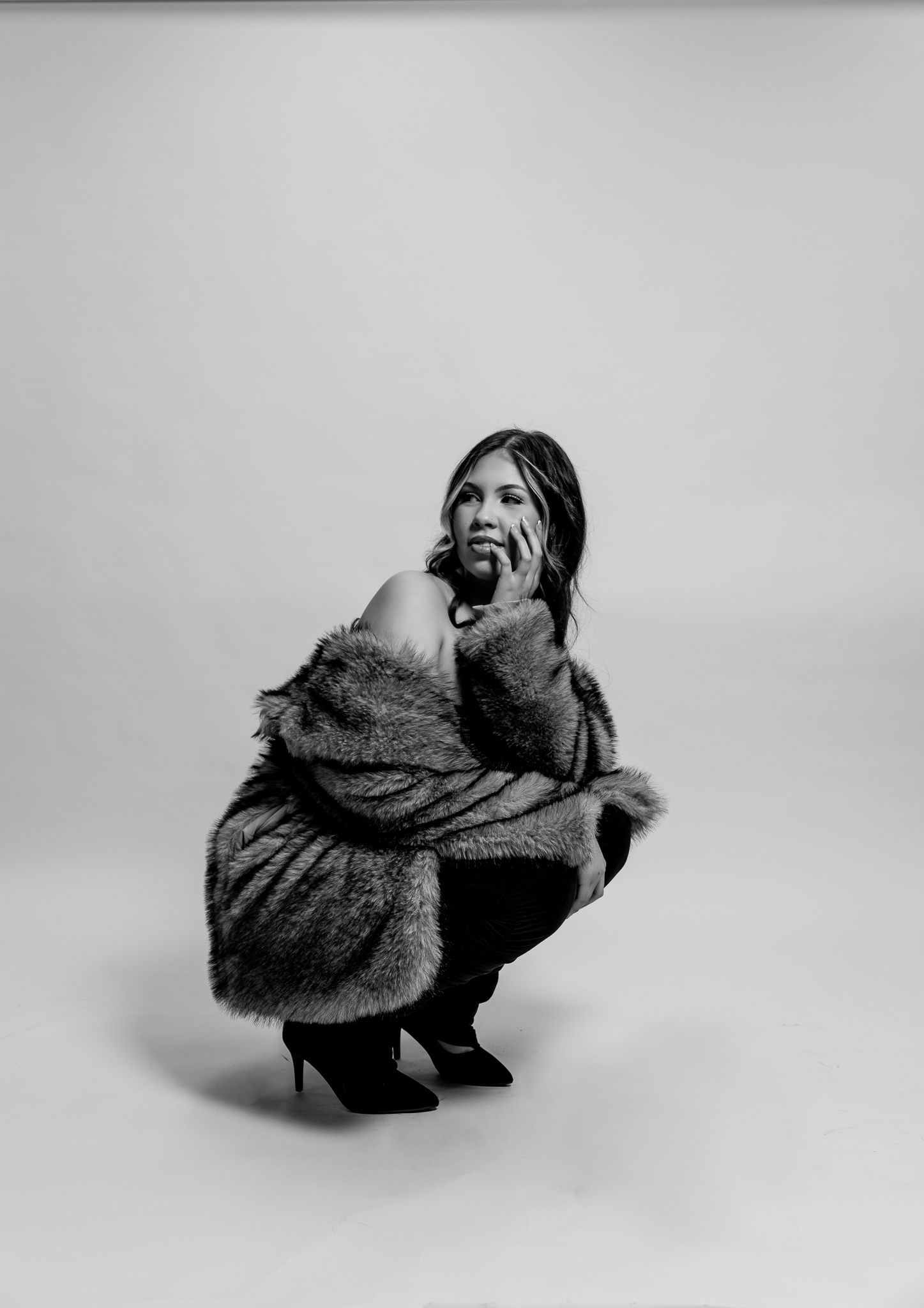Black and white fashion photograph of a person in a cozy fur coat crouching in a dramatic pose against neutral backdrop.