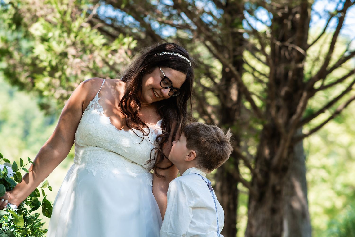 A tender moment shared between two people in white attire outdoors under sunlit trees.