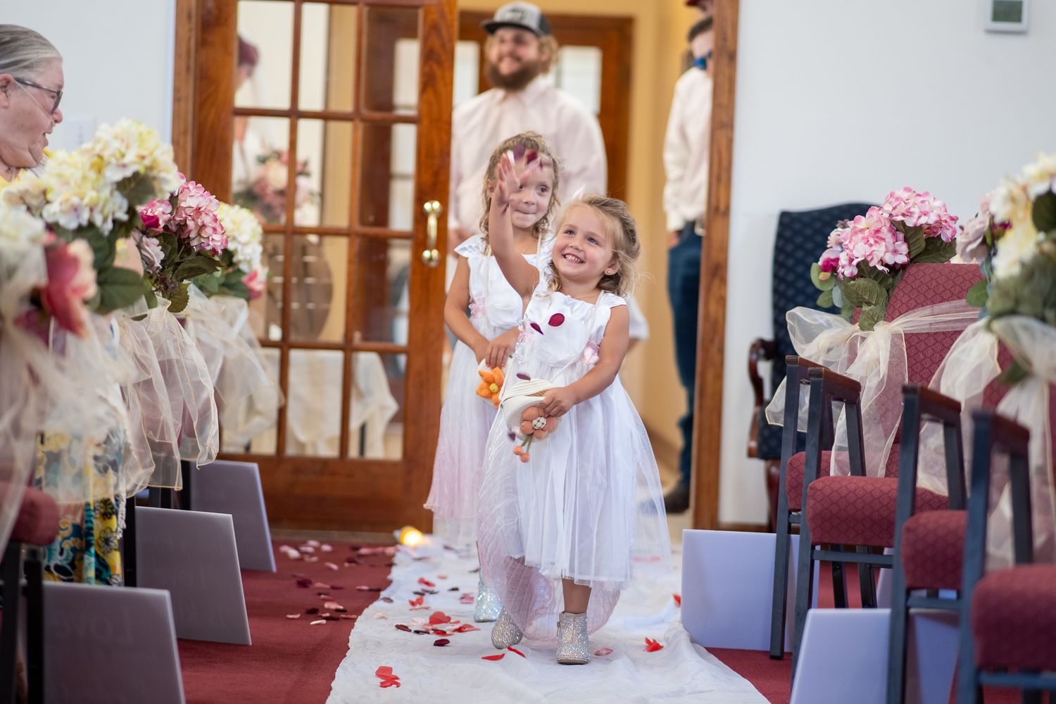 Two small children in white dresses stand on a rose petal-covered aisle runner during a wedding ceremony.