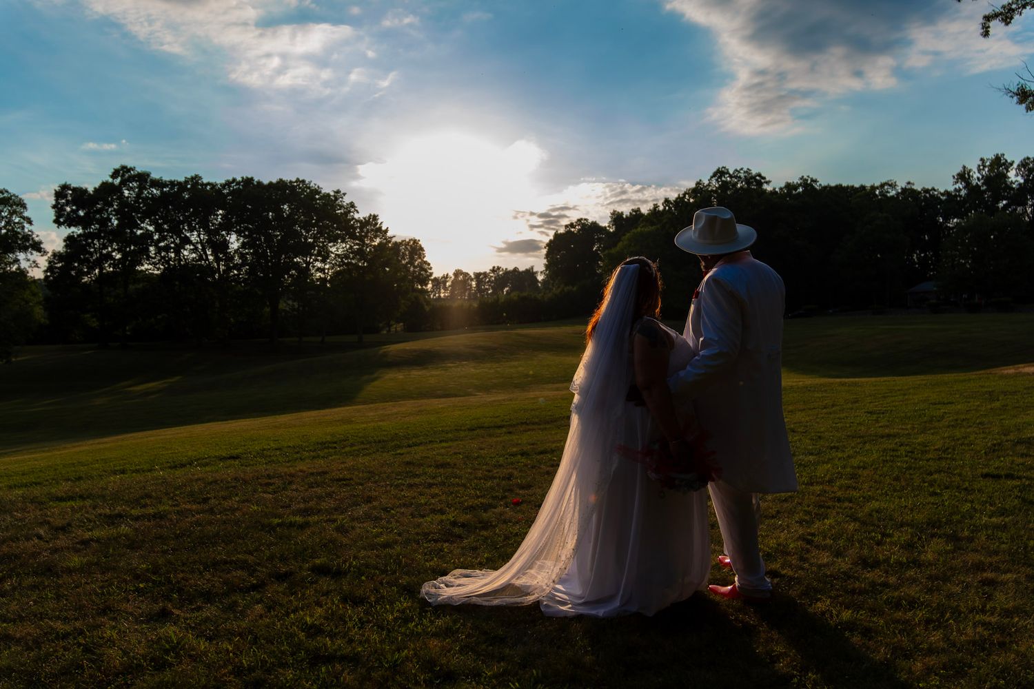Silhouetted couple embraces in a grassy field at sunset with trees on the horizon.