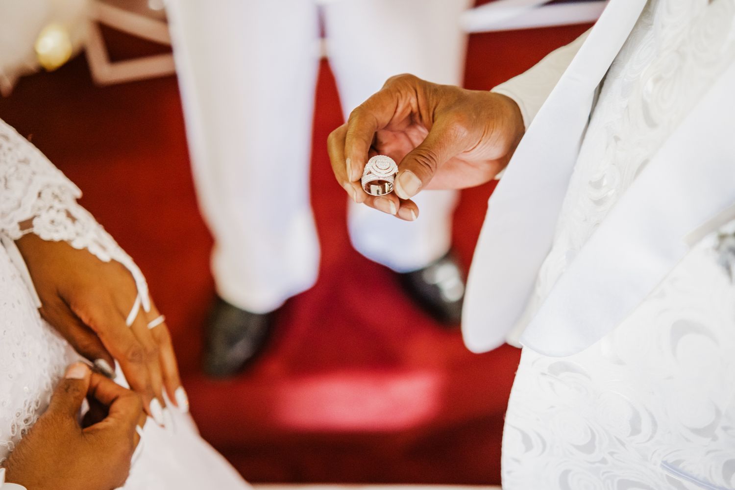 Small brown dog rests on red carpet between figures wearing white clothing on wedding day.