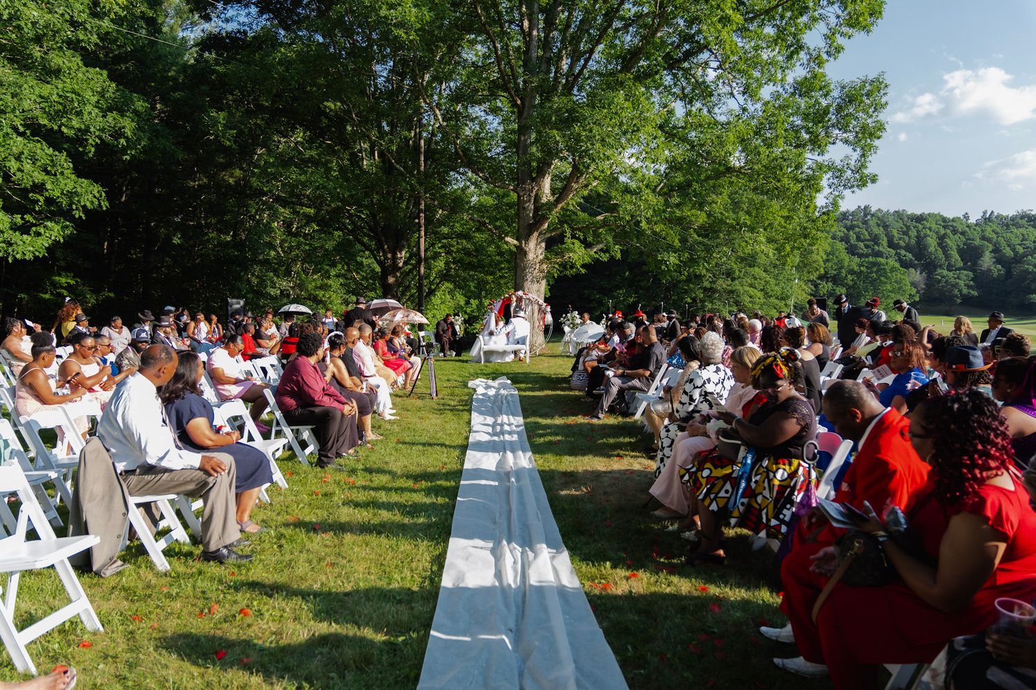 Outdoor wedding ceremony setup with rows of white chairs and red accents along a white aisle runner.