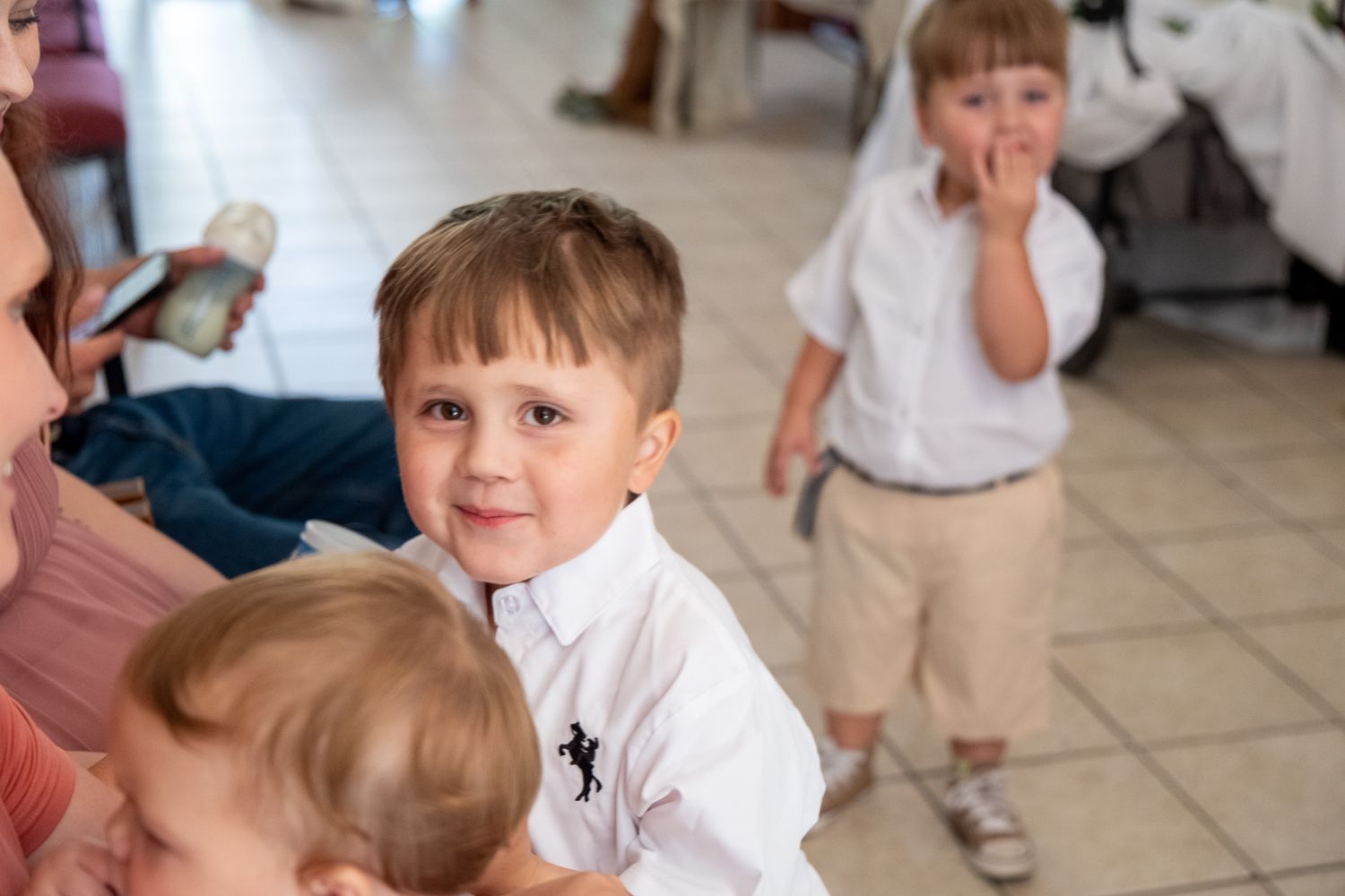 Children in white polo shirts play and make silly faces at an indoor event on a tiled floor.