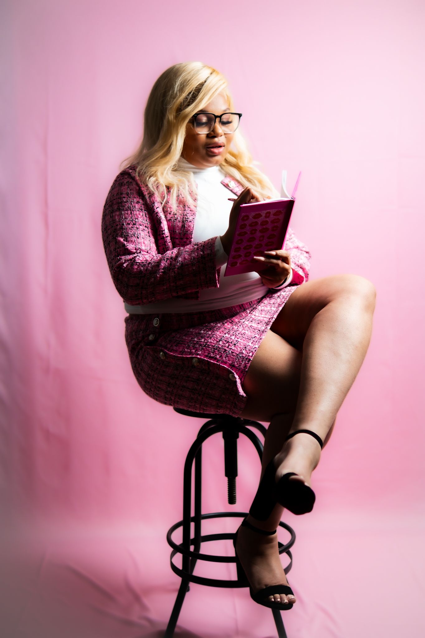 Person in pink cardigan reading book while sitting on stool against pink background.