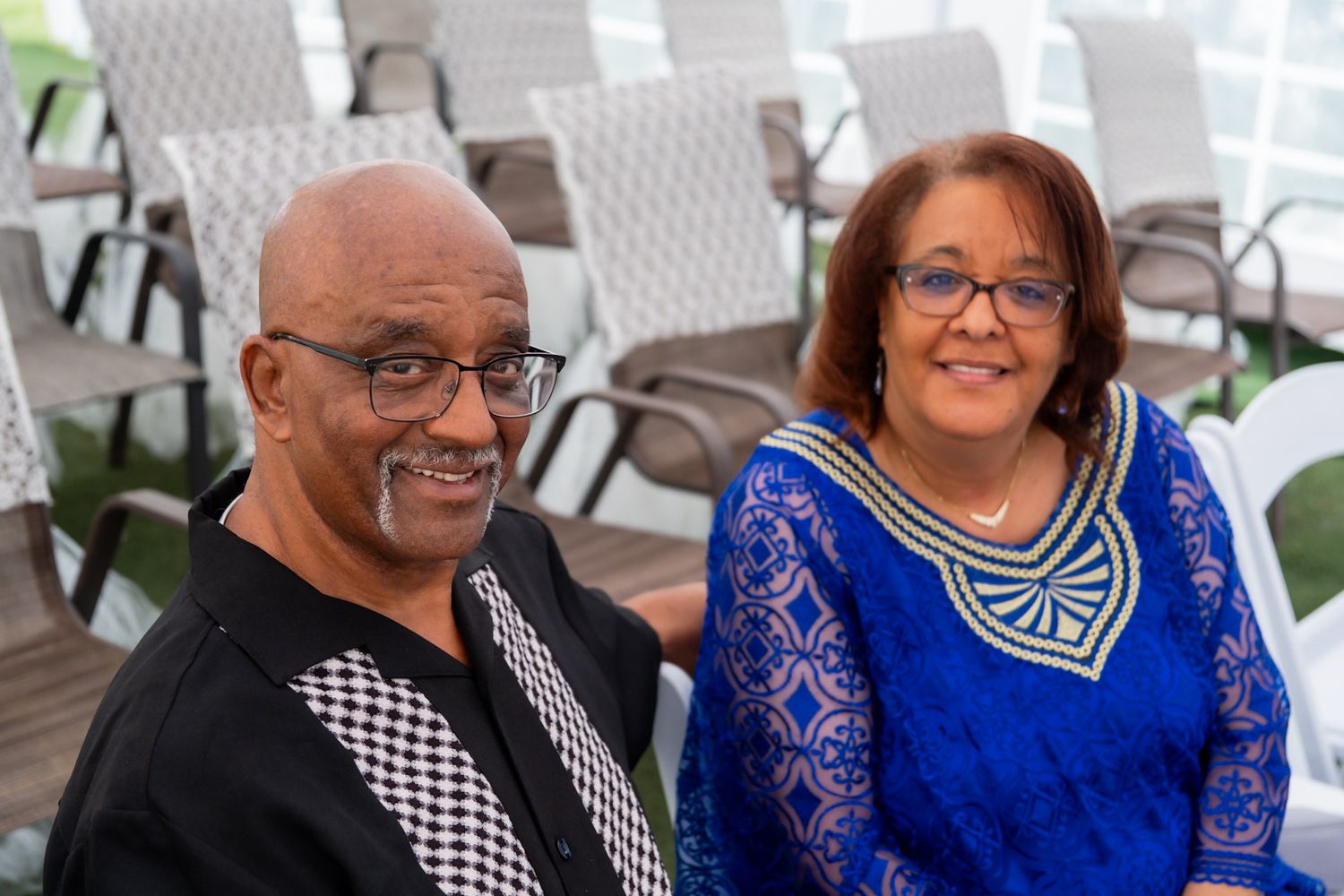 A couple in formal attire sitting together at an event with rows of white chairs behind them.