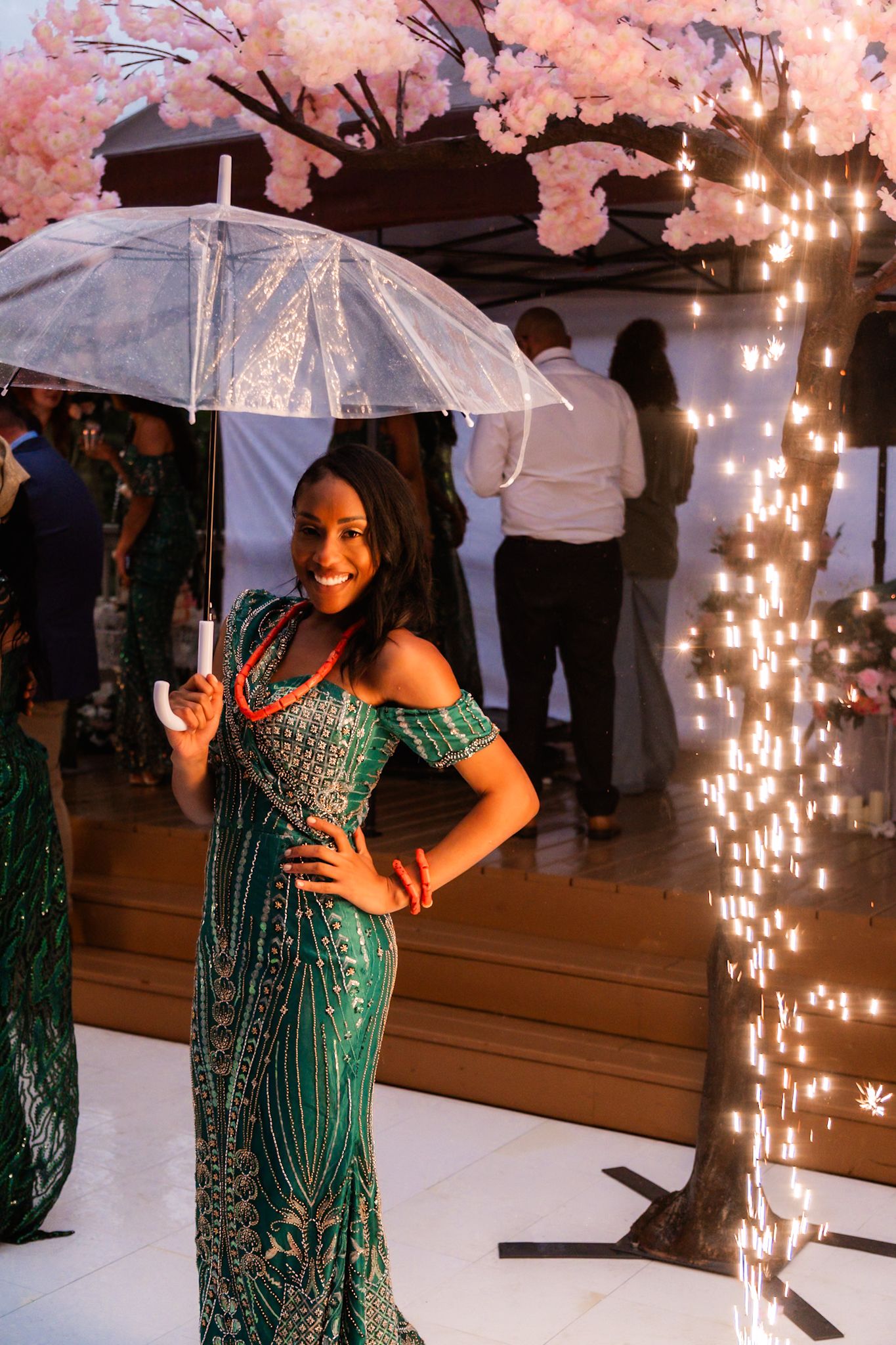 A guest in a sparkly green dress poses with a clear umbrella under cherry blossoms and twinkle lights.