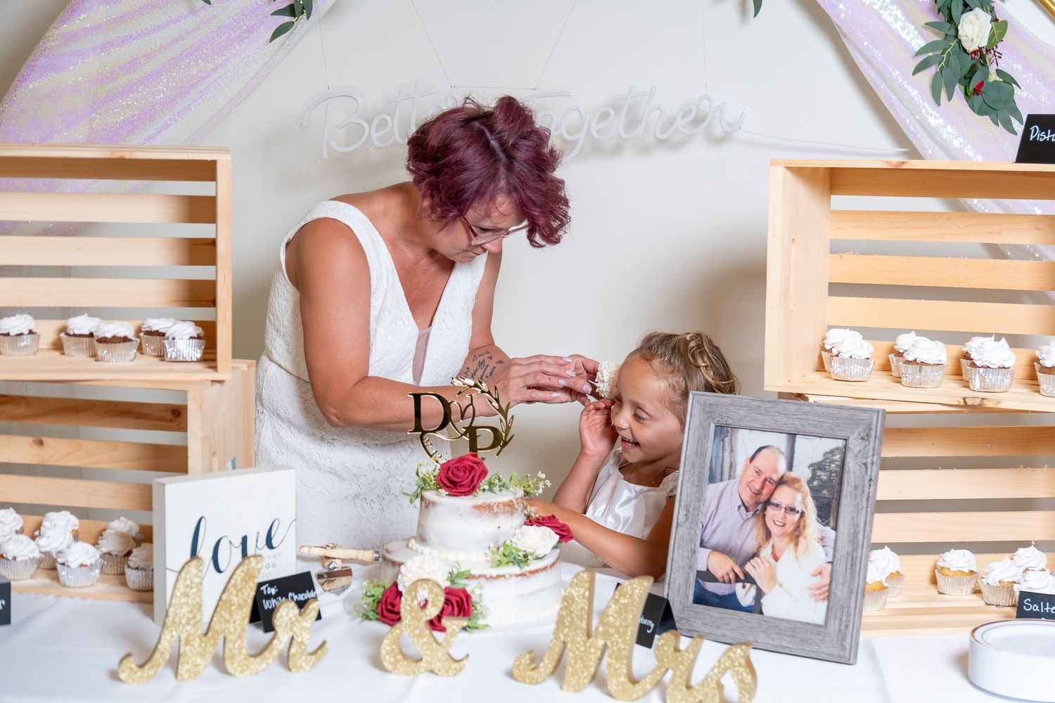 Two people arrange desserts and decorative items on a white table display with wooden shelving and floral accents.