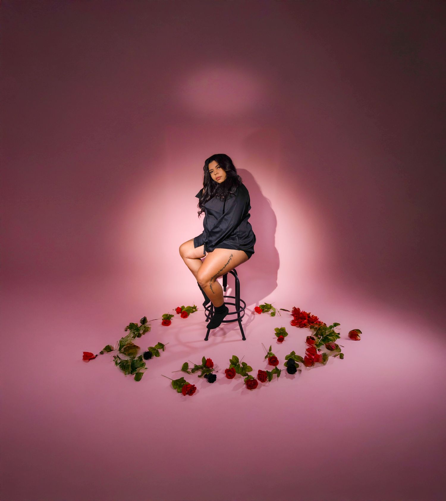 Dramatic studio portrait on stool surrounded by red roses against pink backdrop.