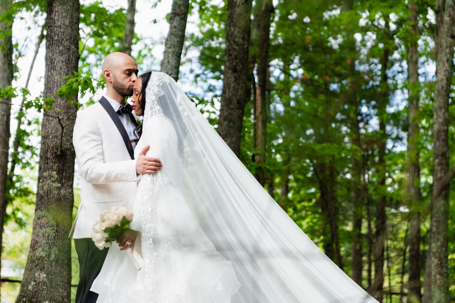 Wedding couple in white attire posing among trees in woodland setting.