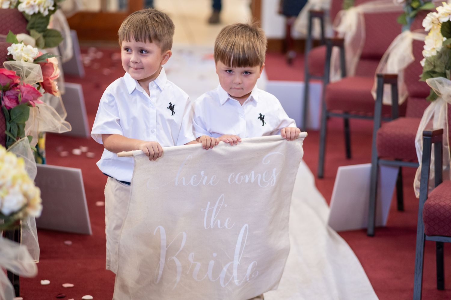 Children walk down a church aisle carrying signs at a wedding ceremony with red carpeted floors and wooden pews.