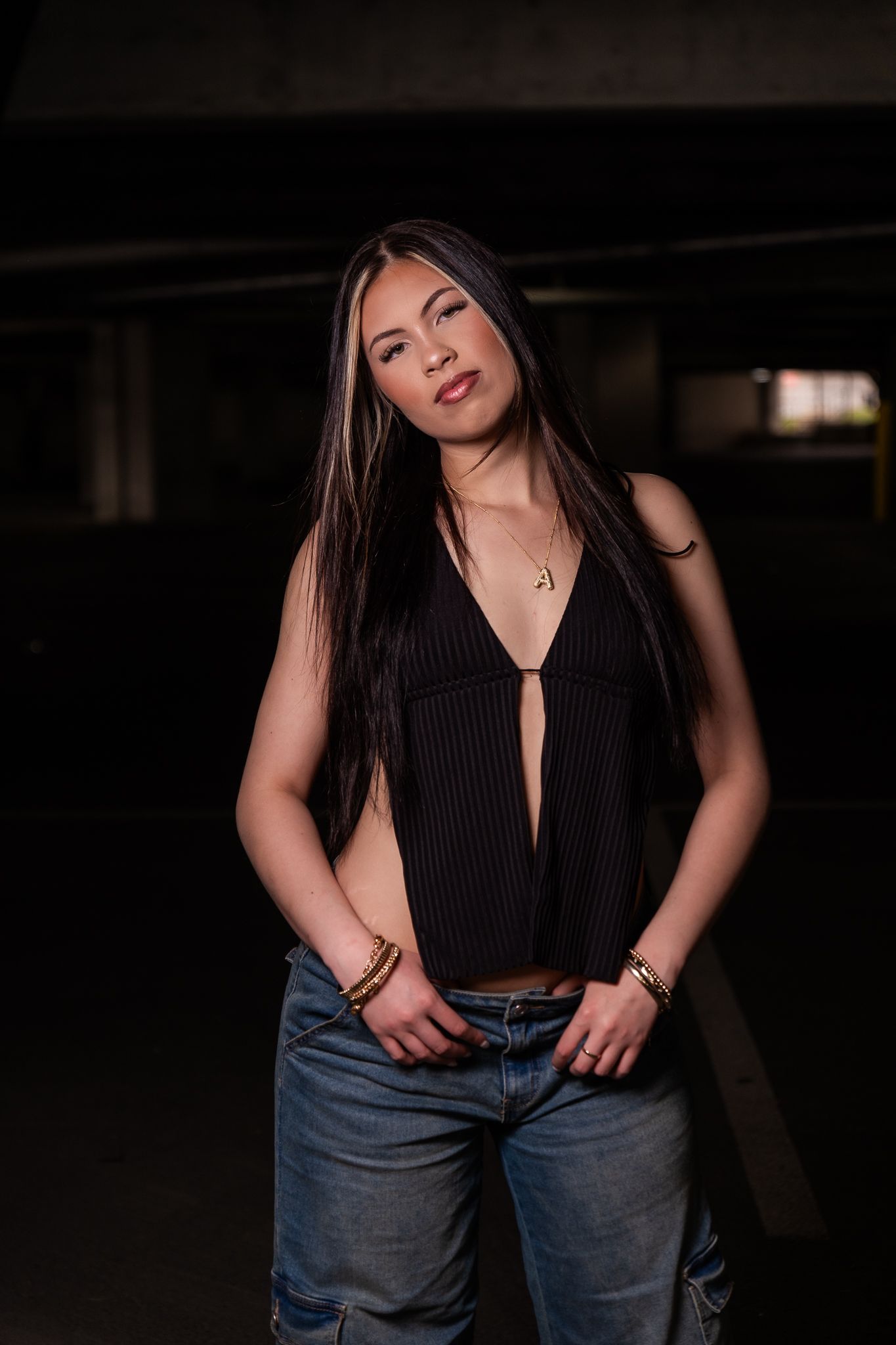 Moody portrait of a woman in a dark pinstripe top and jeans posing in a dimly lit parking structure.