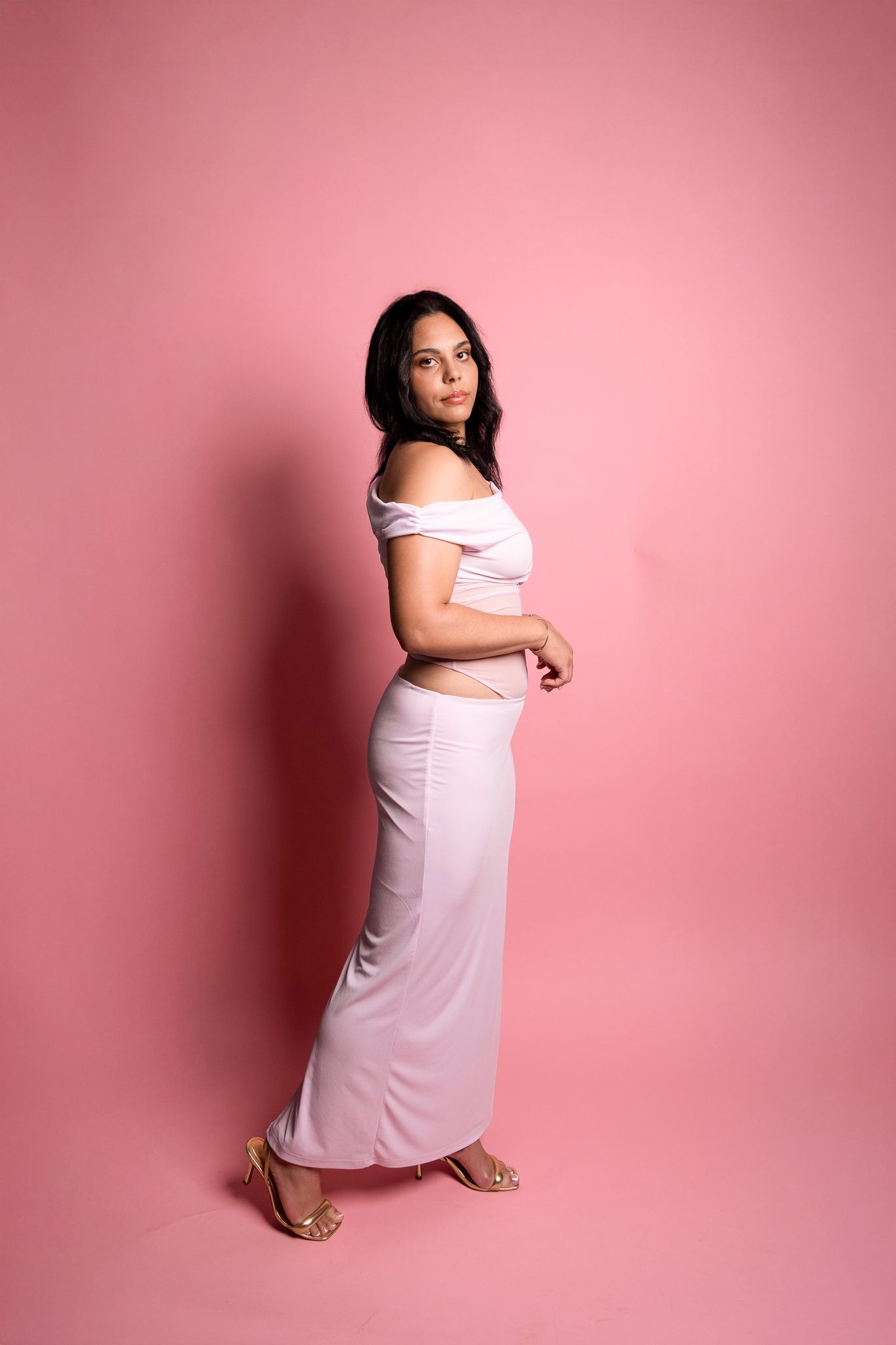 A pregnant woman in a white dress poses against a pink backdrop during a maternity photoshoot.