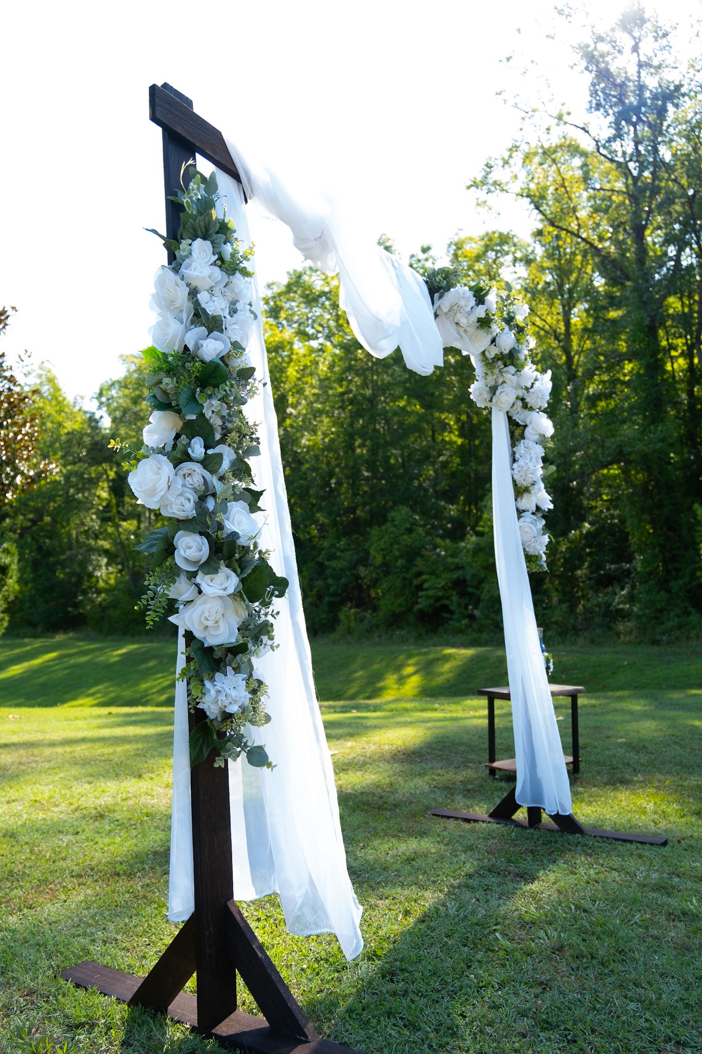 A wedding arch decorated with white flowers and draped fabric stands in a sunlit garden.