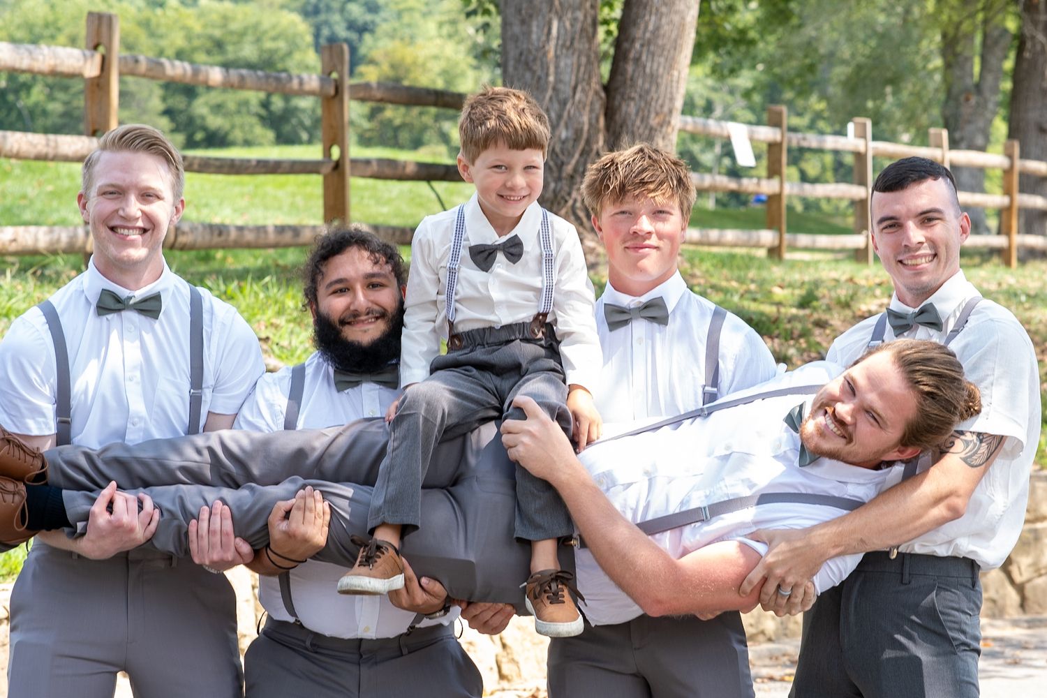 Groomsmen in matching suspenders and bowties playfully carrying someone at an outdoor venue.