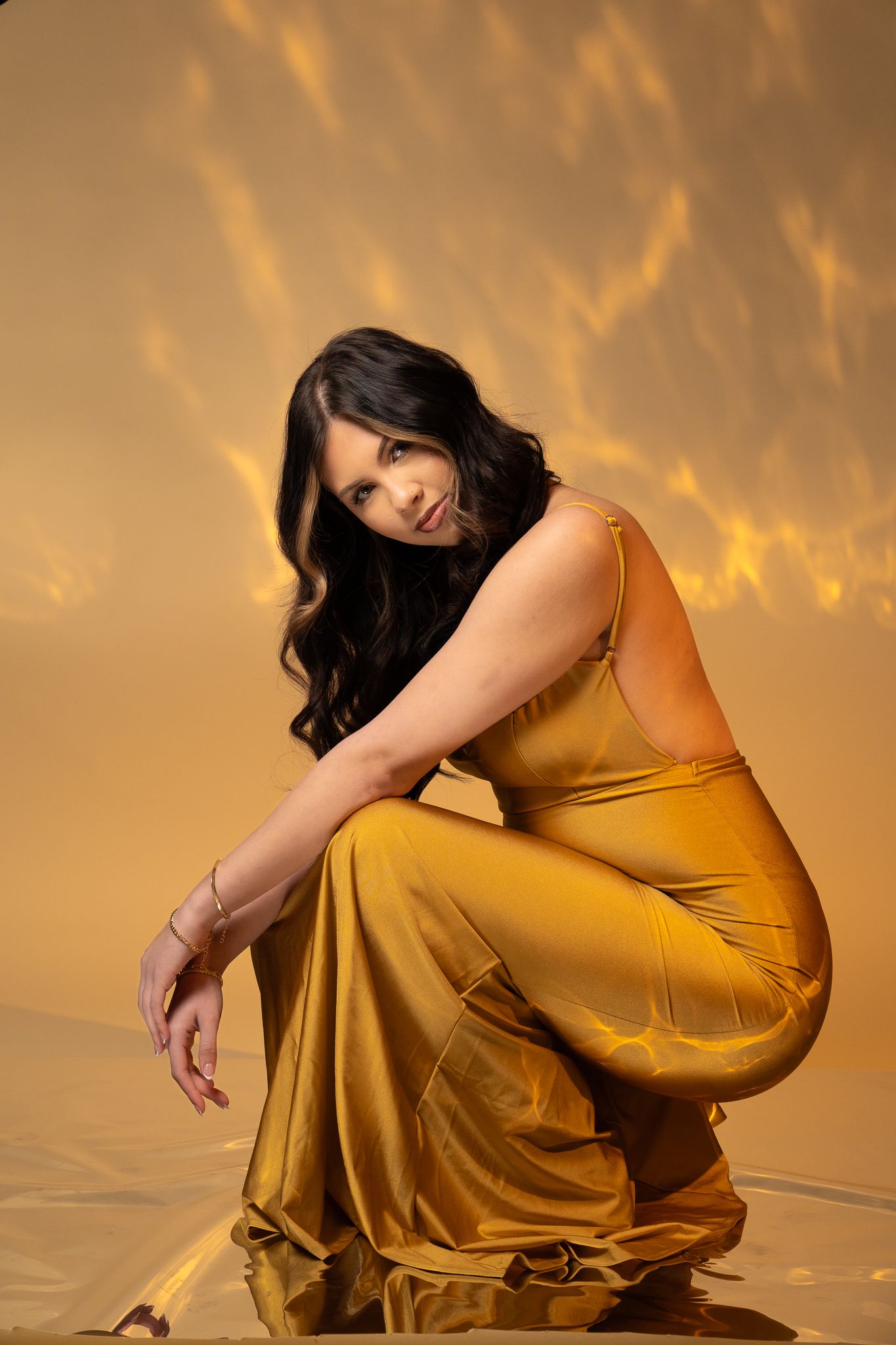 Woman in flowing mustard yellow dress poses elegantly against golden backdrop with dramatic lighting effects.
