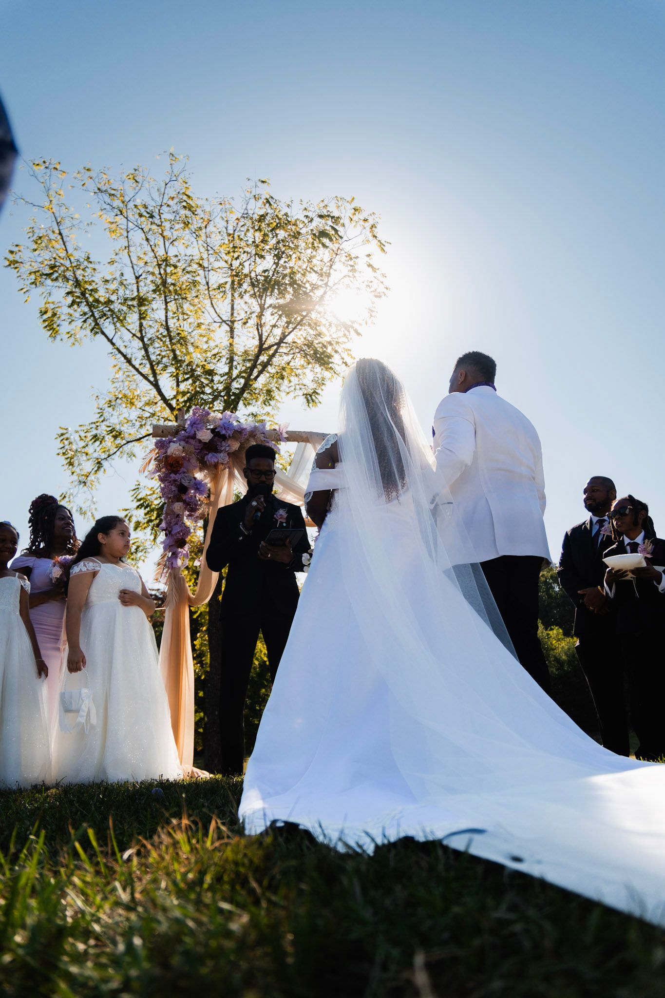 Scenic wedding ceremony under tree with flowing white dress in foreground.