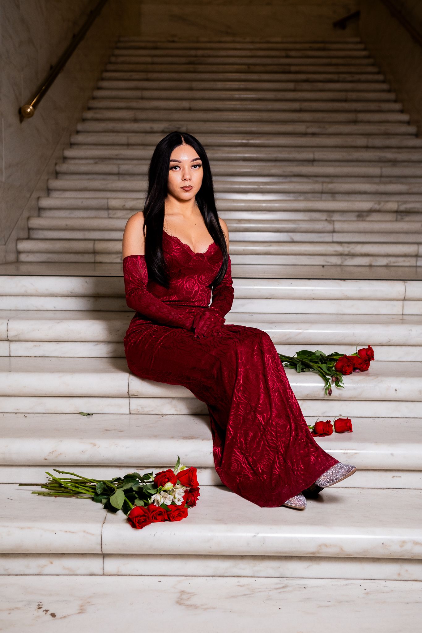 Elegant portrait in red lace gown on marble staircase with scattered roses.
