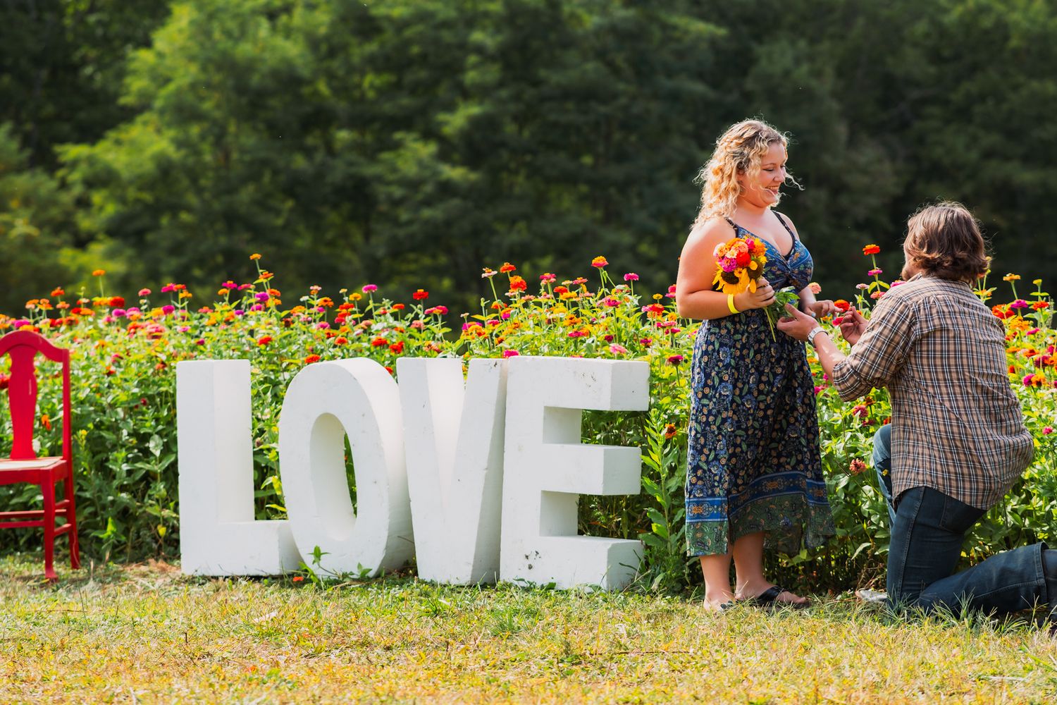 A proposal moment captured in a flower garden with white LOVE letters and a red bench in the background.