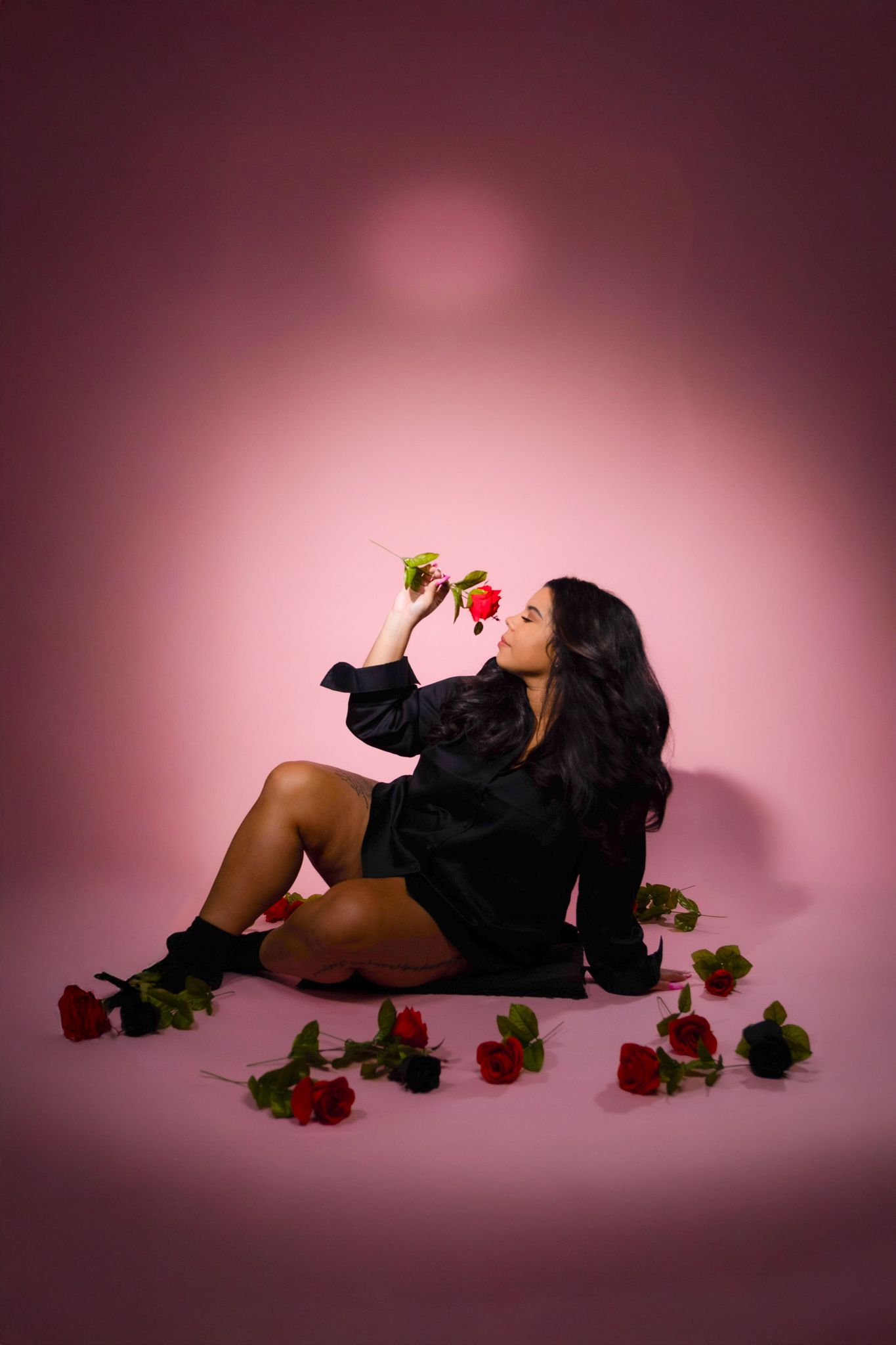 Artistic photo of a person in black attire posing with red roses against a pink backdrop.