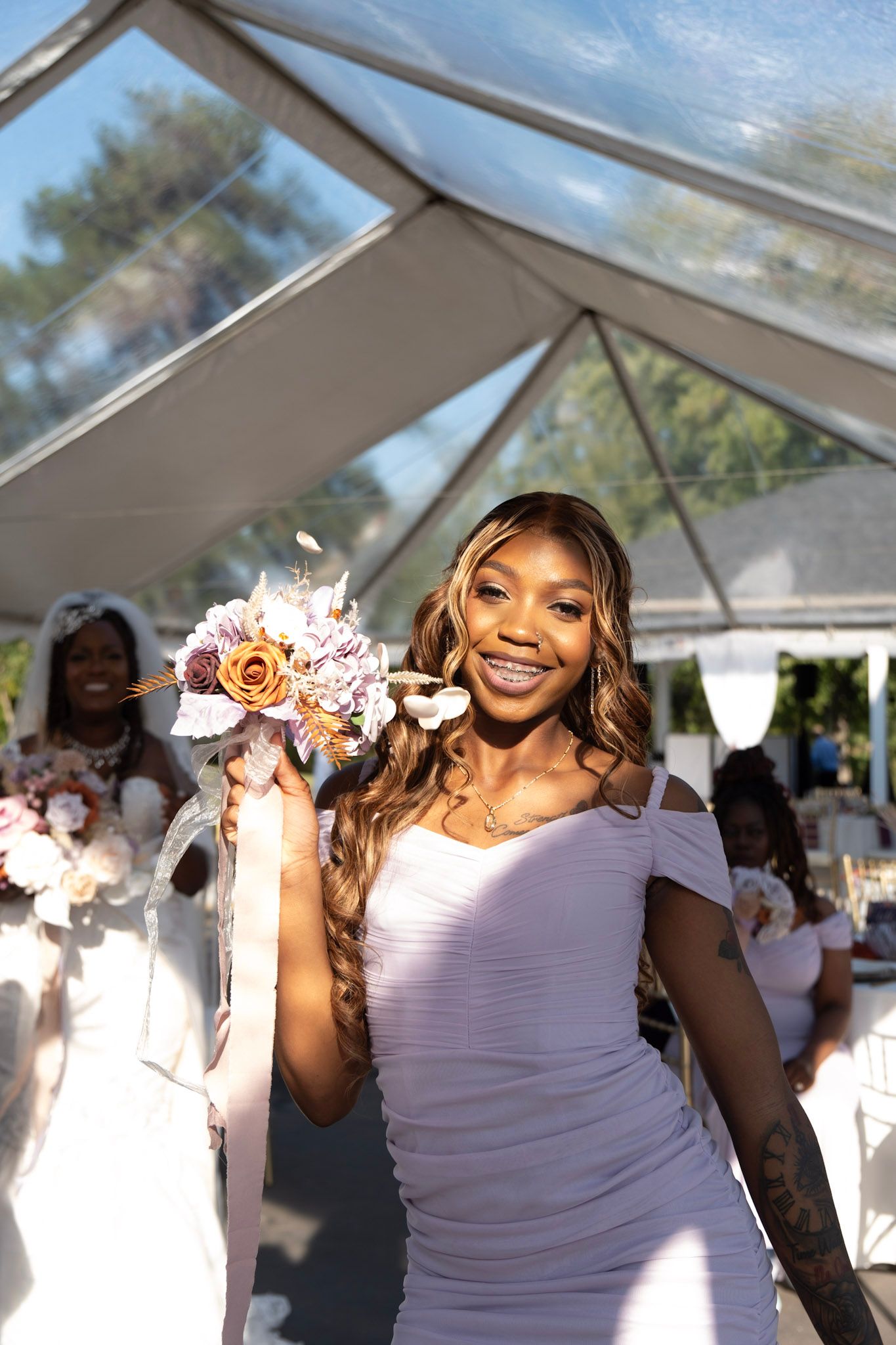 A wedding attendee in a lavender dress holds a pink and peach bouquet under a clear tent.
