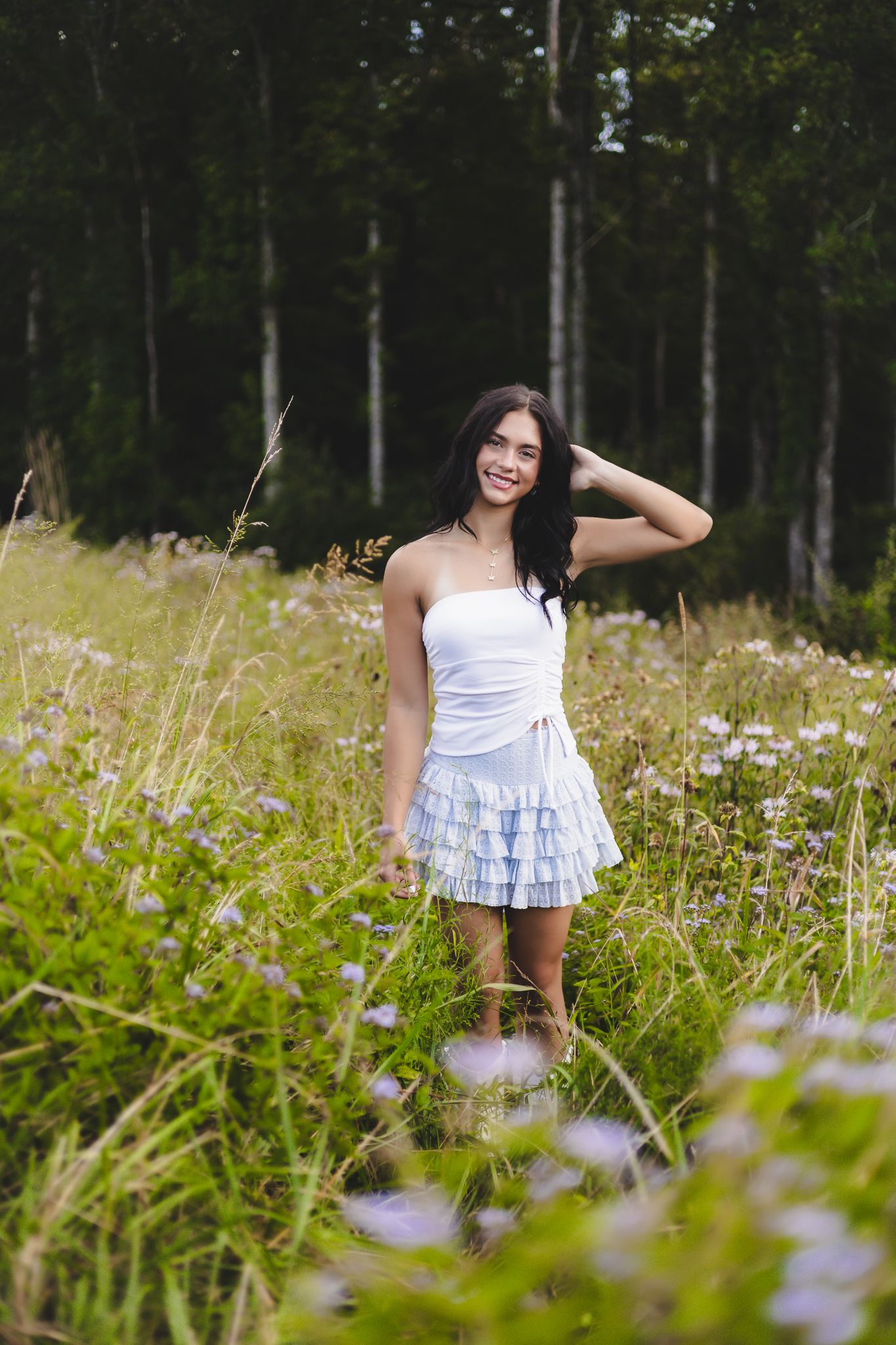 A white-dressed figure stands among purple wildflowers in a sunny meadow with trees in the background.