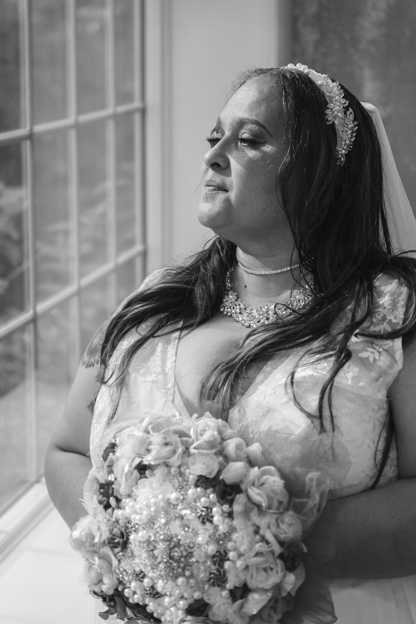 Black and white portrait of a bride holding a bouquet while looking out a window.