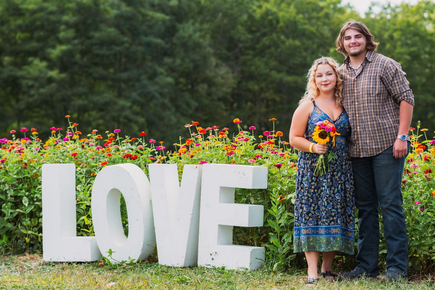 A couple poses smiling beside large white LOVE letters in a blooming flower garden.