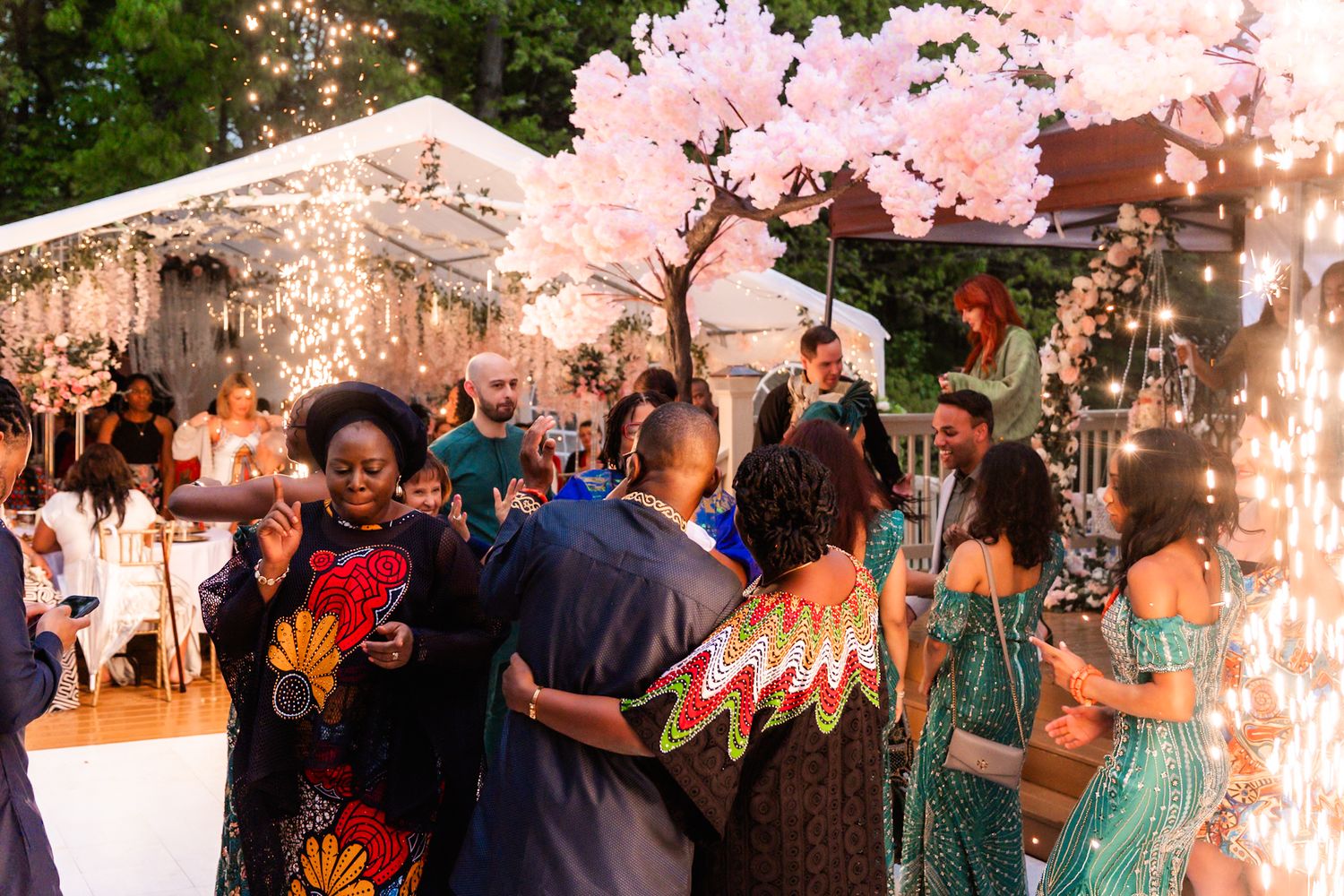 Wedding guests mingle at an outdoor reception under pink trees and string lights.
