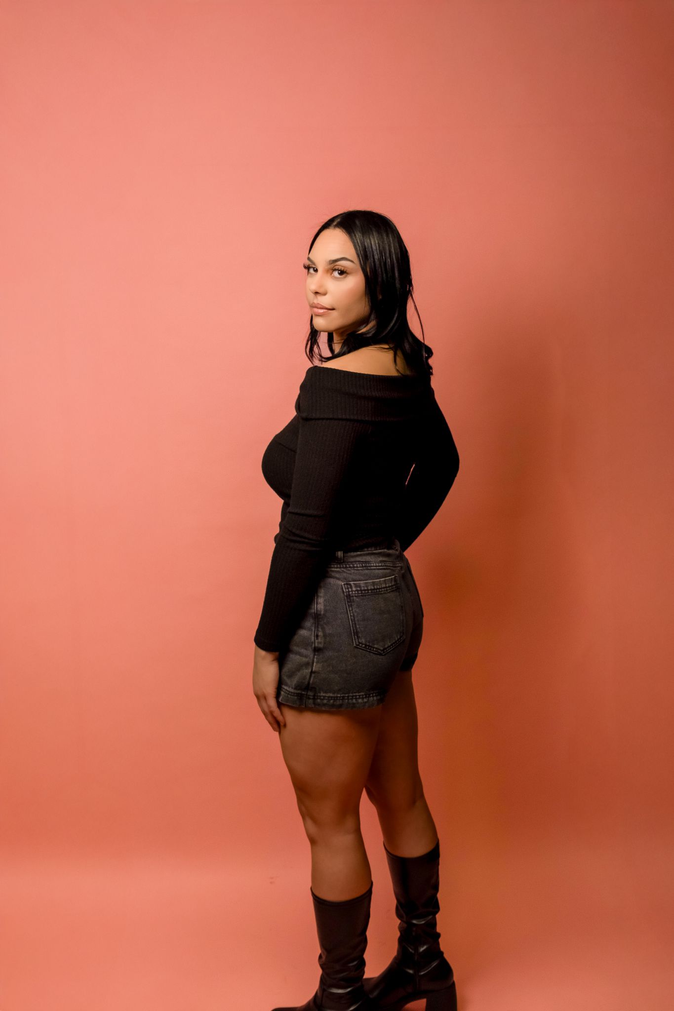 A figure in a black off-shoulder top and denim shorts poses confidently against a coral-colored backdrop.