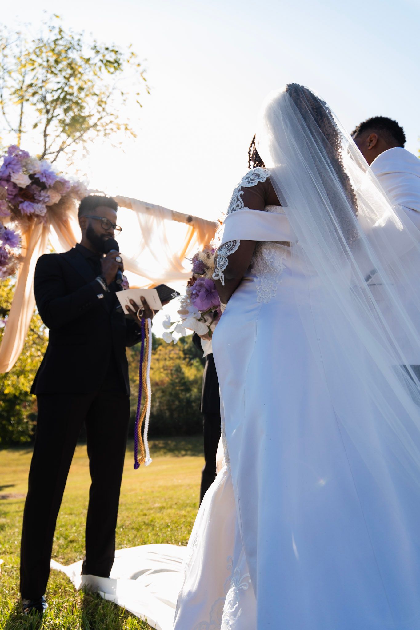 A wedding ceremony takes place outdoors on a sunny day with purple floral arrangements.