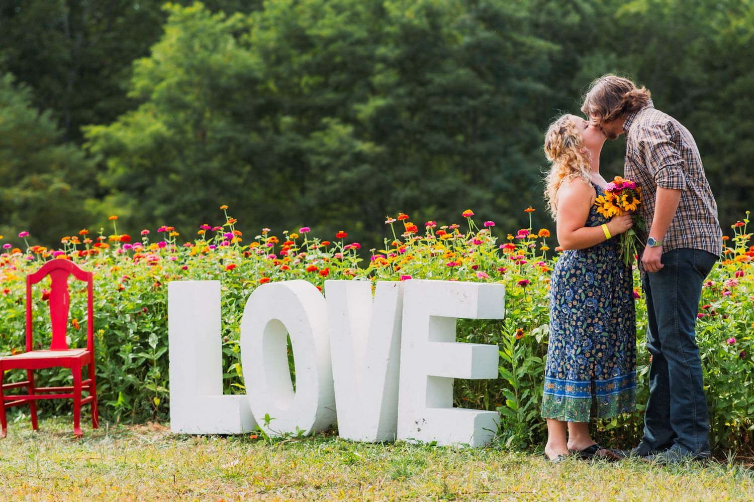 A couple shares an intimate moment in front of large white LOVE letters in a garden filled with red flowers.