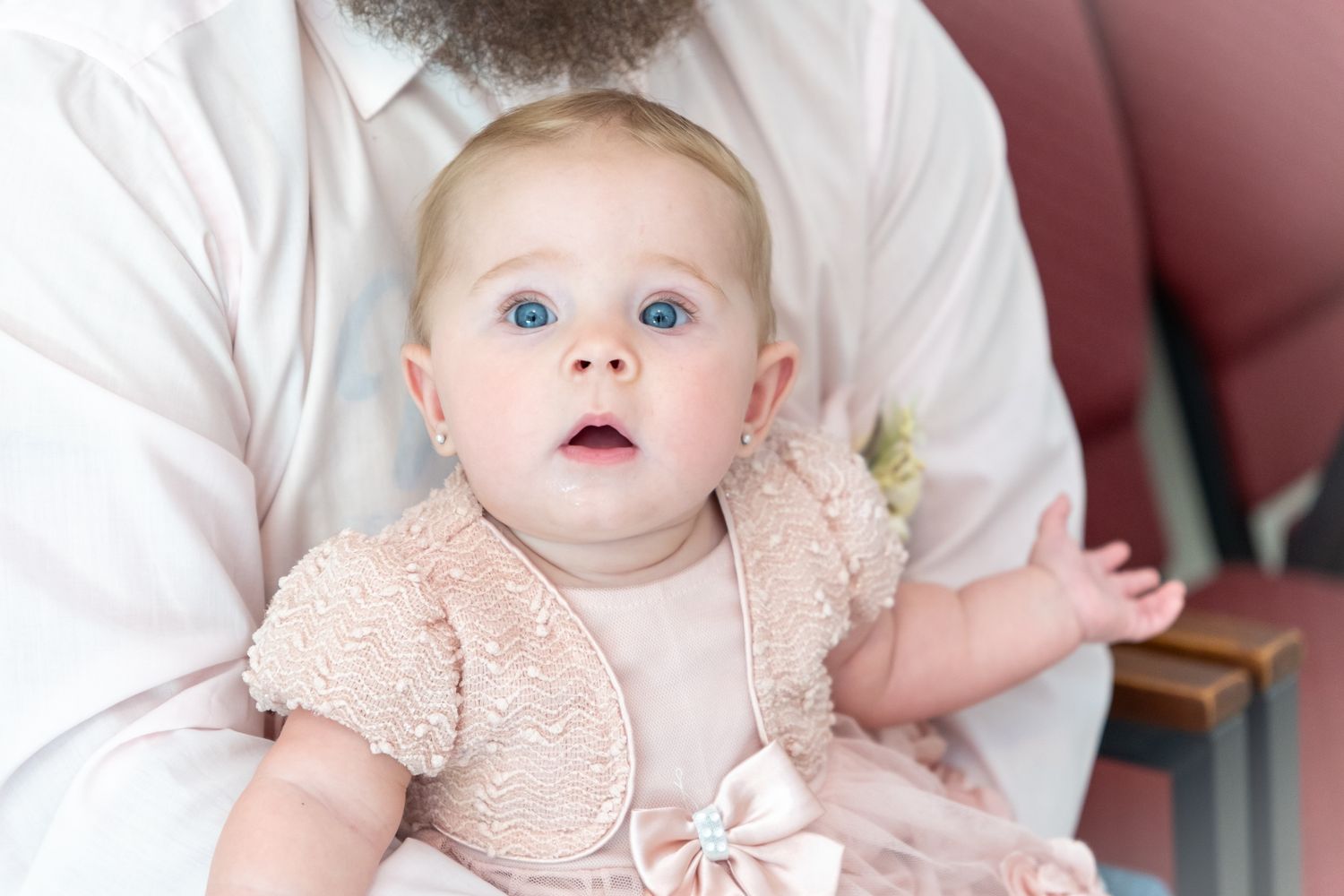 Baby wearing a peach dress with bow details sitting on a plush chair looking upward with an adorable expression.