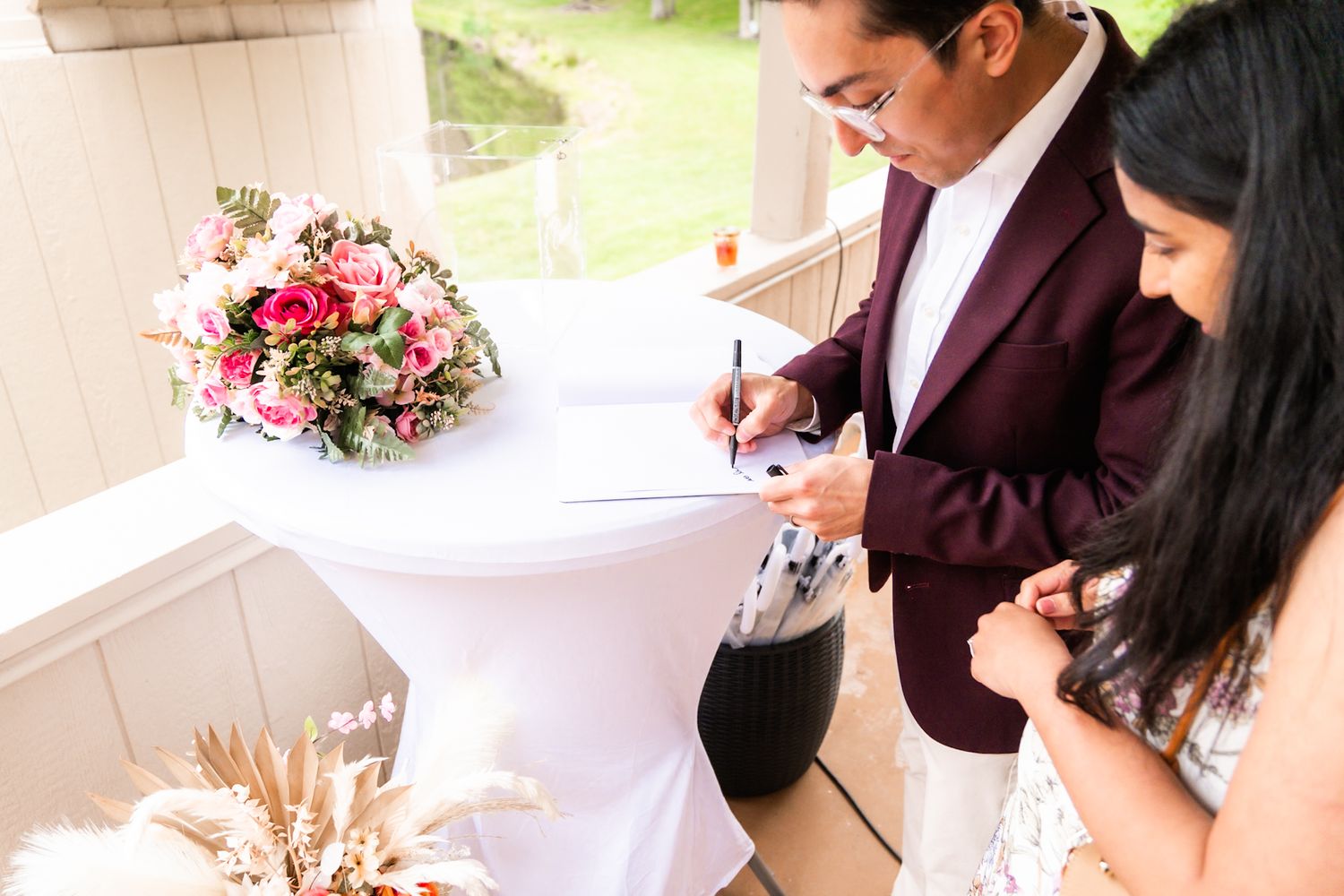 A couple signs documents at a wedding table decorated with a pink floral centerpiece.