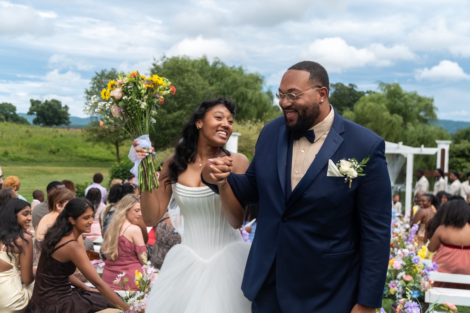 Happy newlywed couple laughing together at outdoor wedding ceremony, bride holding colorful bouquet.