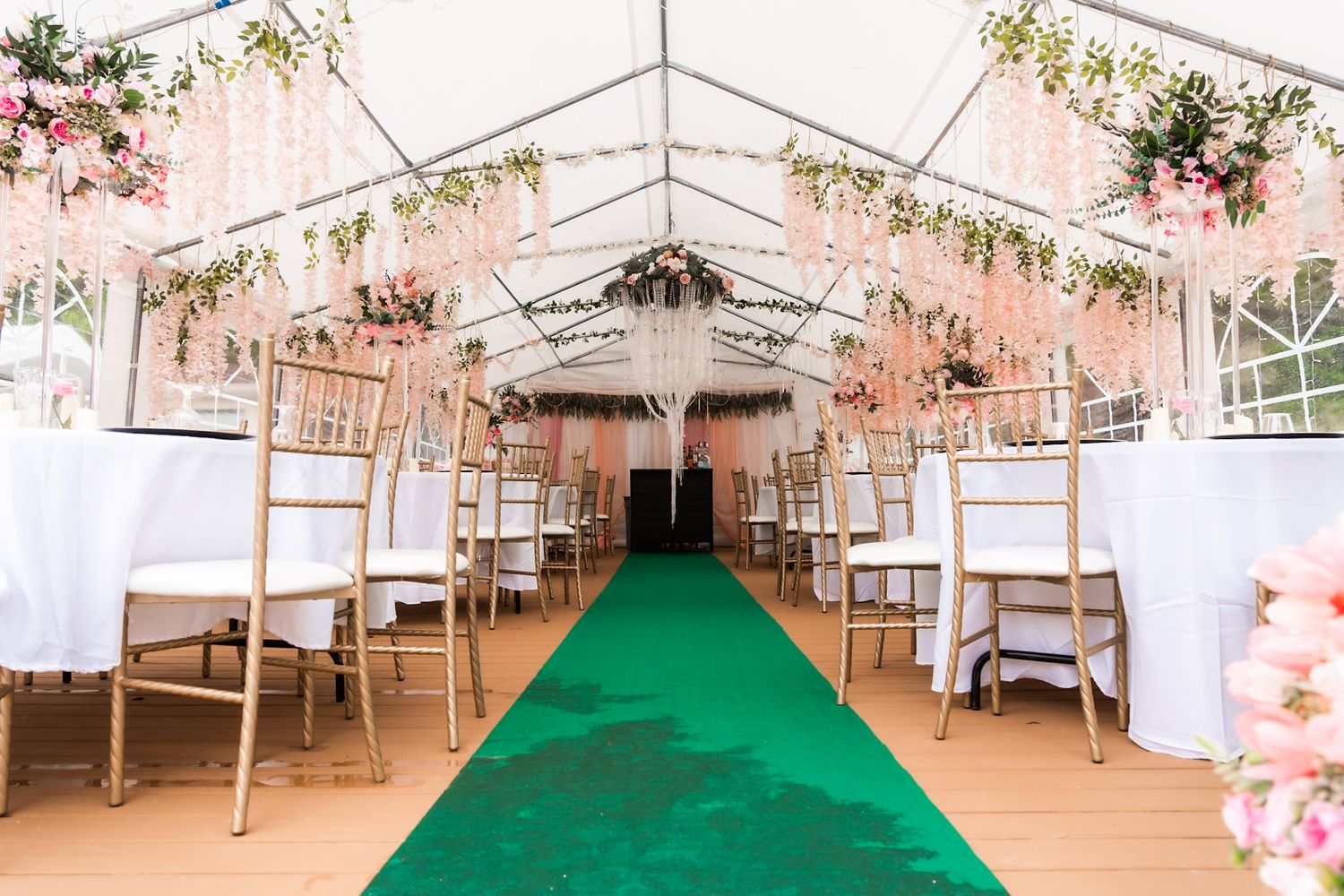 Wedding ceremony tent with green carpet aisle runner and white chairs decorated with pink floral accents.