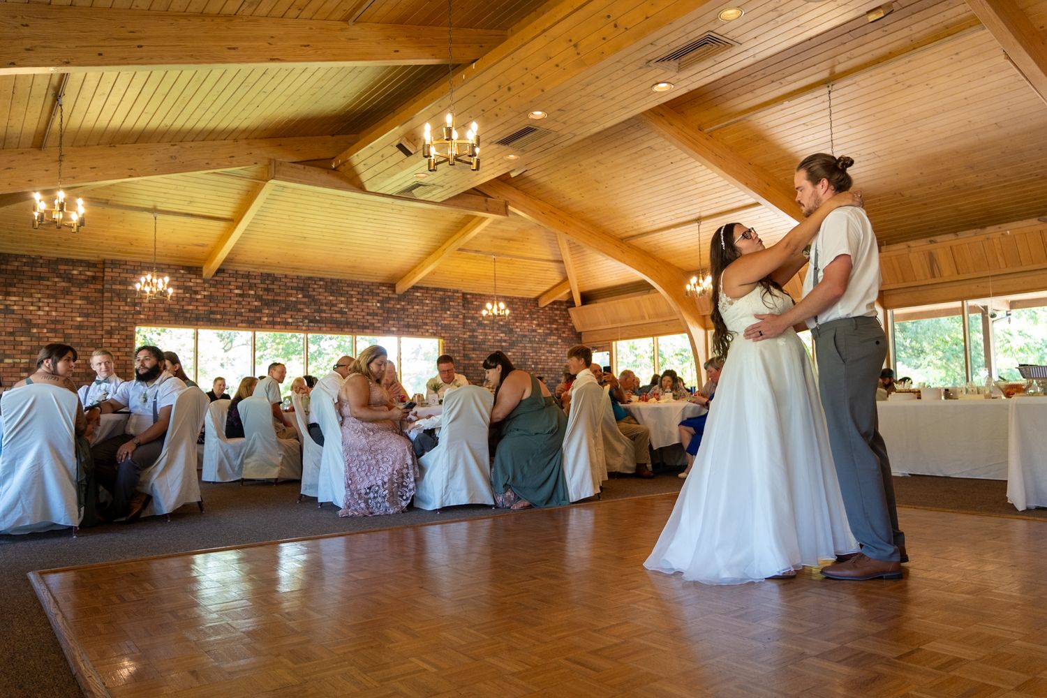 A couple shares their first dance in a rustic wooden reception venue with guests watching.
