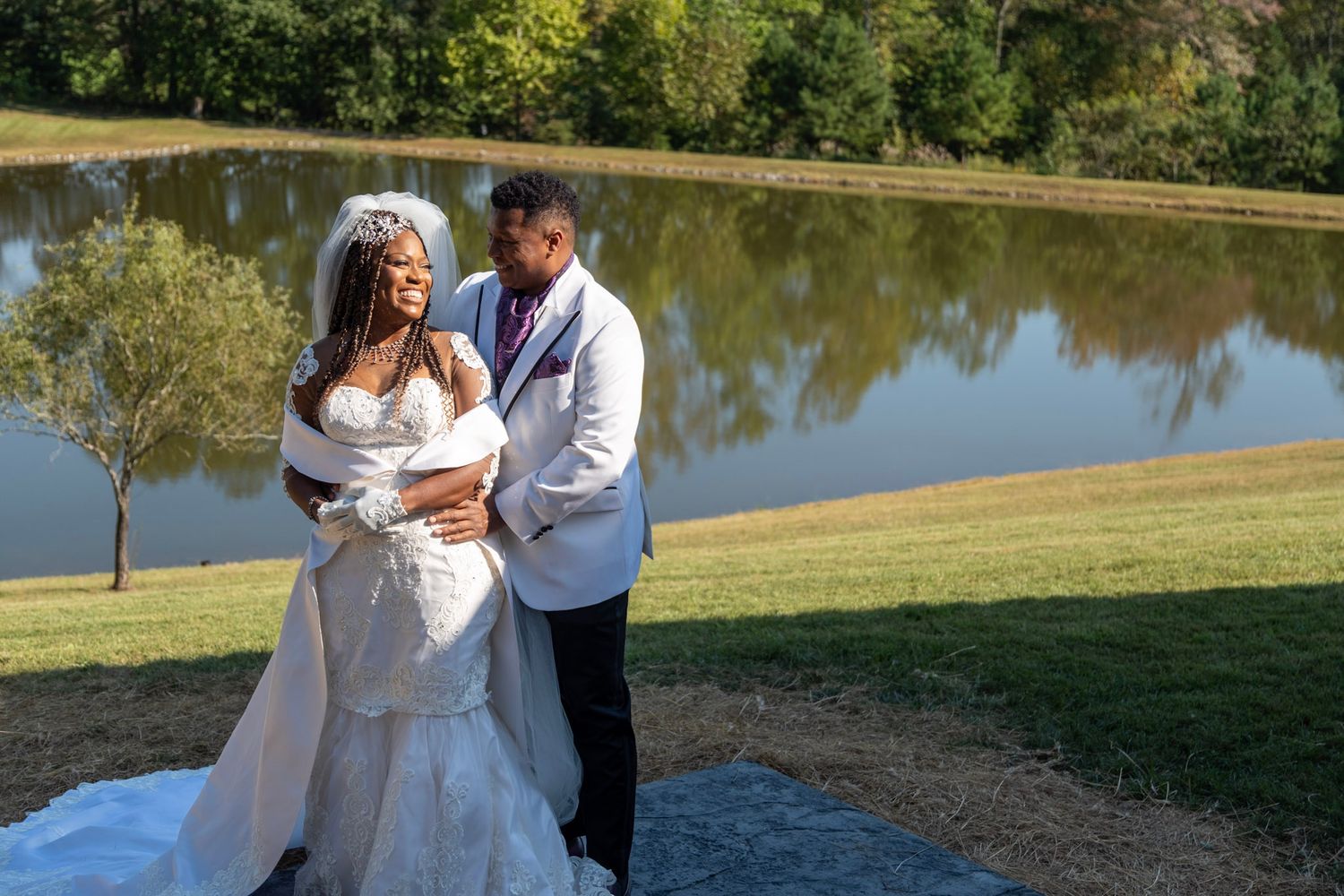 A happy couple embraces by a serene lake on their wedding day, with the bride in white and groom in a light colored jacket.